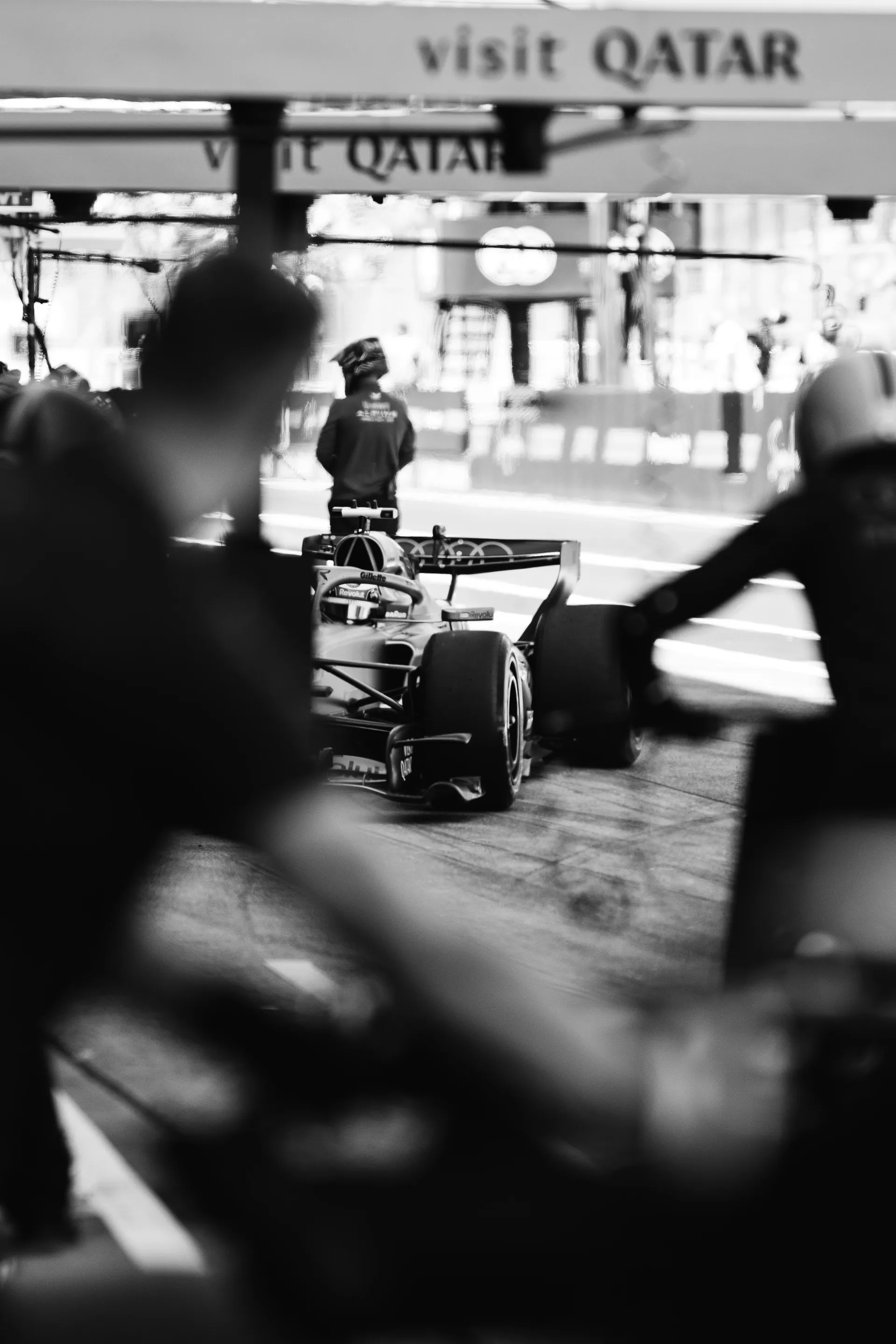 Black and white garage scene with an Audi Revolut F1® Team car positioned near pit lane, blurred crew members in the foreground, and a helmeted team member waiting ahead.