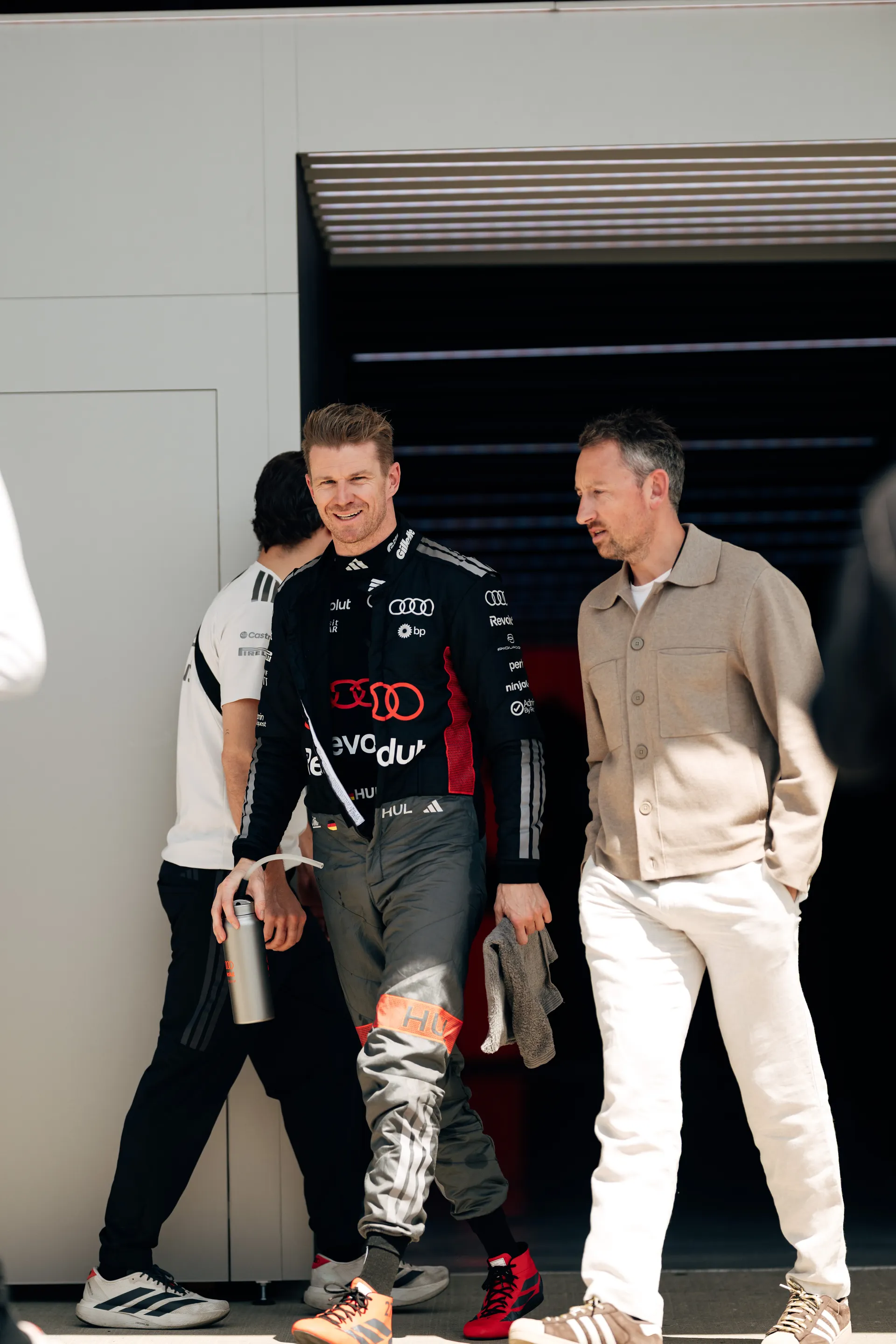 Nico Hulkenberg walks out of the Audi Revolut F1® Team garage in race suit alongside another team member in a beige jacket, while a third person stands partly turned away in the background.