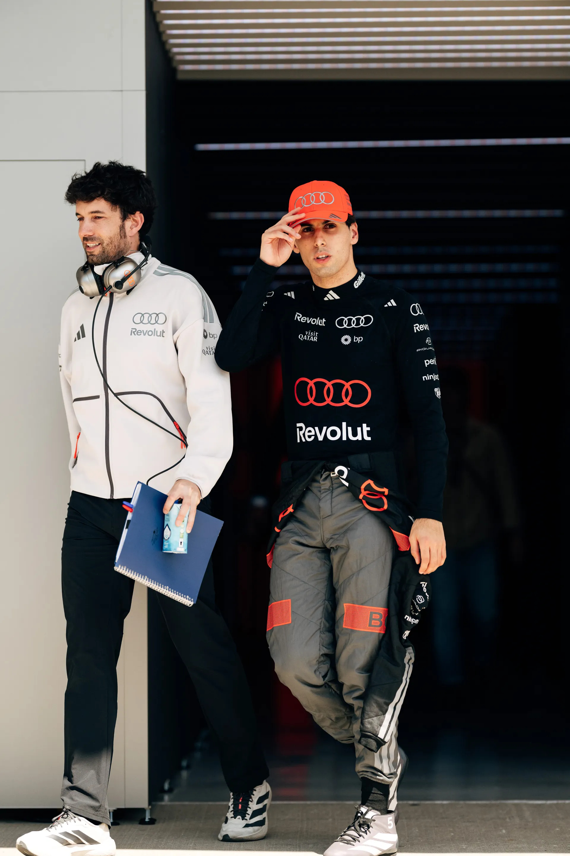 Color image of Gabriel Bortoleto Mini stepping out of the garage in Audi Revolut F1® Team kit and orange cap beside a team member carrying a notebook and wearing a headset.