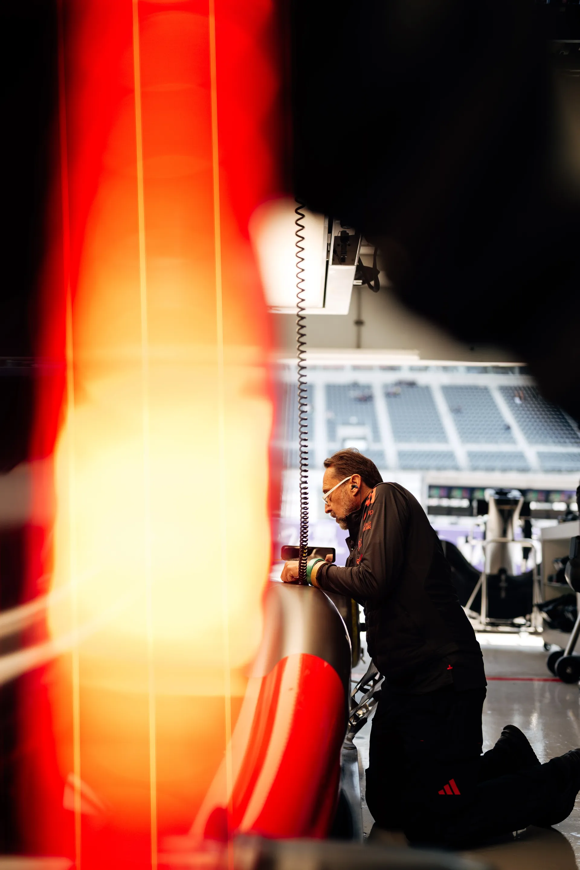 Team member kneeling beside the side of the Audi Revolut F1® Team car in the garage, framed by a red light flare and the blurred grandstand outside.