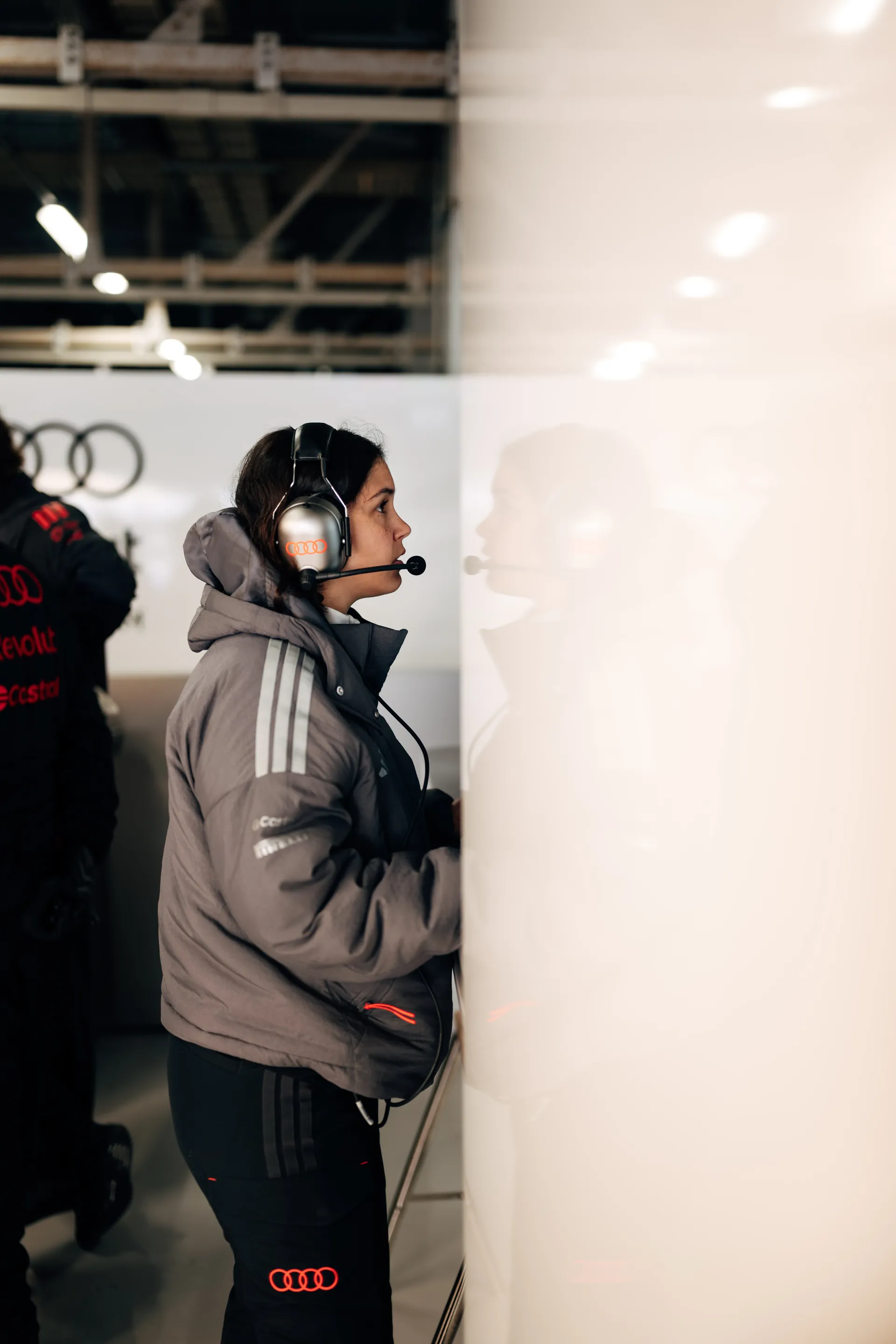 Female Audi Revolut F1® Team engineer wearing a headset and standing side on in the garage, focused on the wall and her reflection.