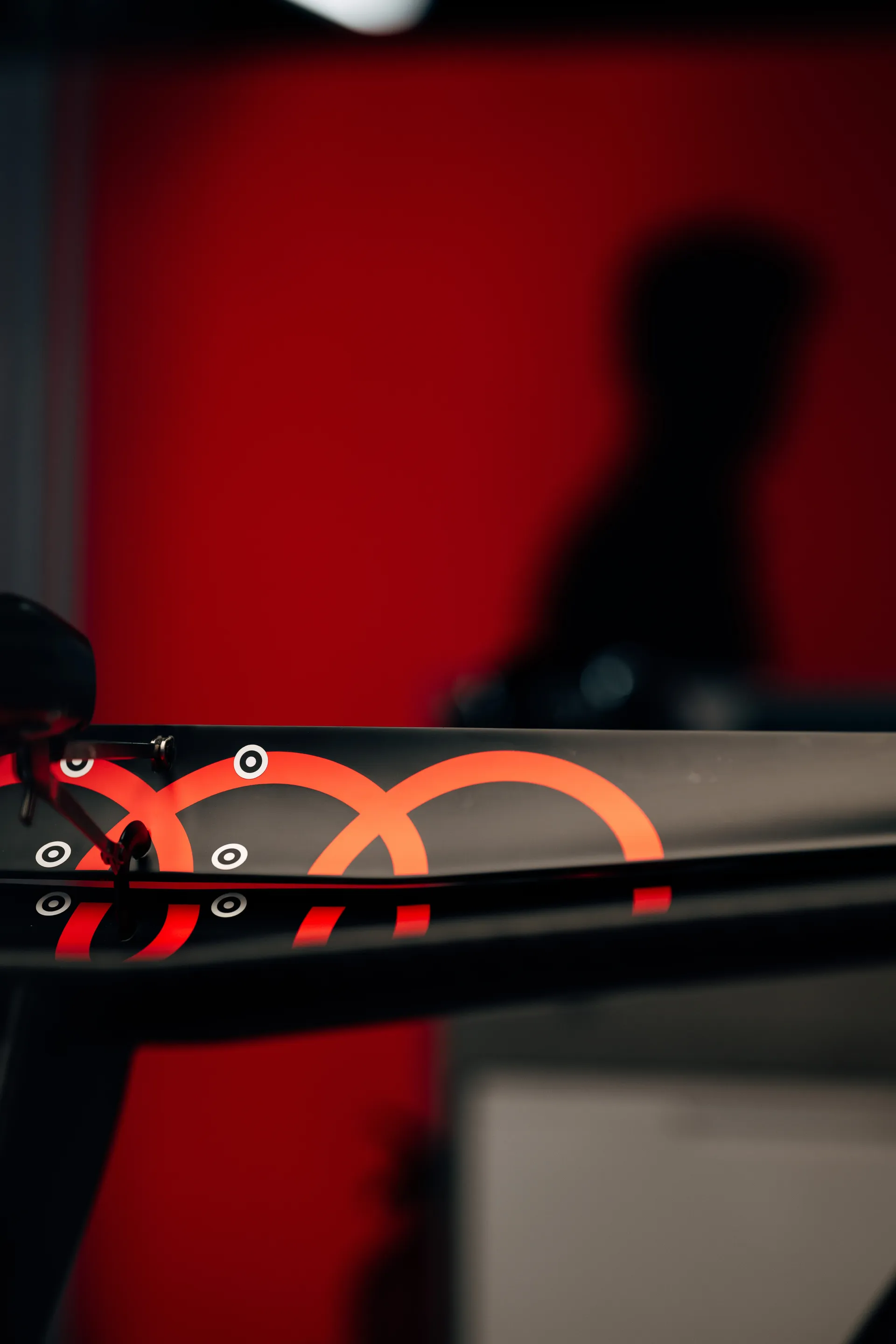 Detail of the Audi Revolut F1® Team rear wing with glowing red Audi rings, set against a red wall and a blurred team member silhouette.