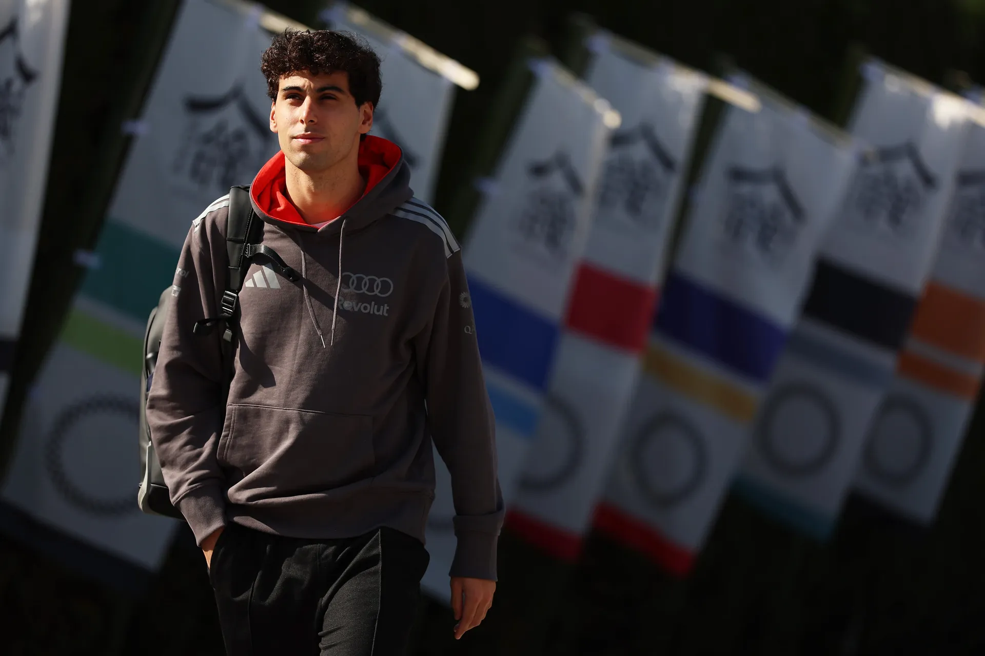 Gabriel Bortoleto walks outdoors in Audi Revolut F1® Team travelwear with a backpack over one shoulder, passing a row of colorful Suzuka Circuit banners.