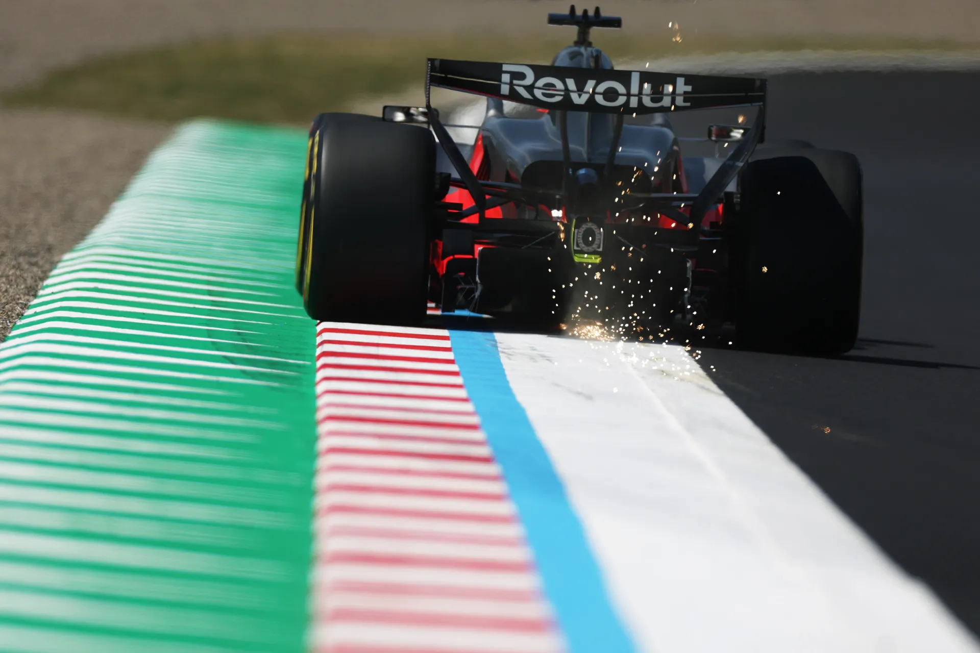 Rear view of an Audi Revolut F1® Team Formula 1 car riding the curb at Suzuka, throwing bright sparks along the green, white, blue, and red track edge.