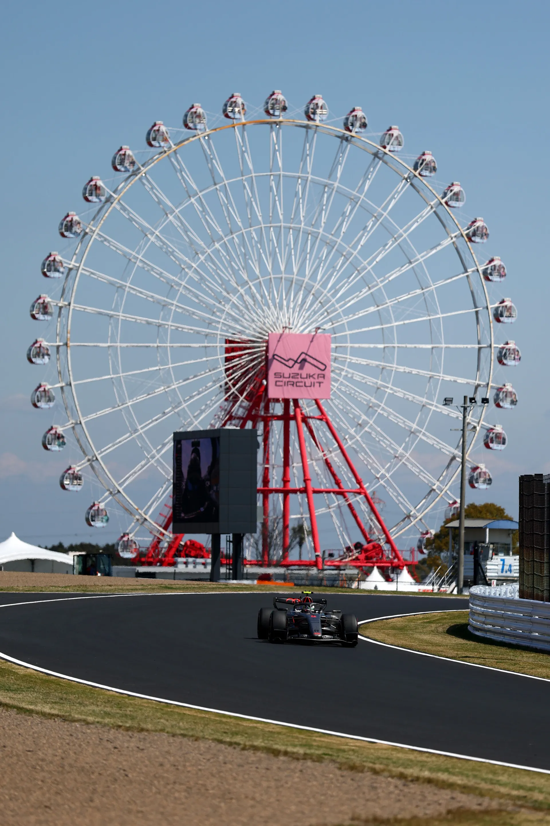 An Audi Revolut F1® Team Formula 1 car drives past the Suzuka Circuit Ferris wheel on a clear day, with the red and white wheel towering above the track.