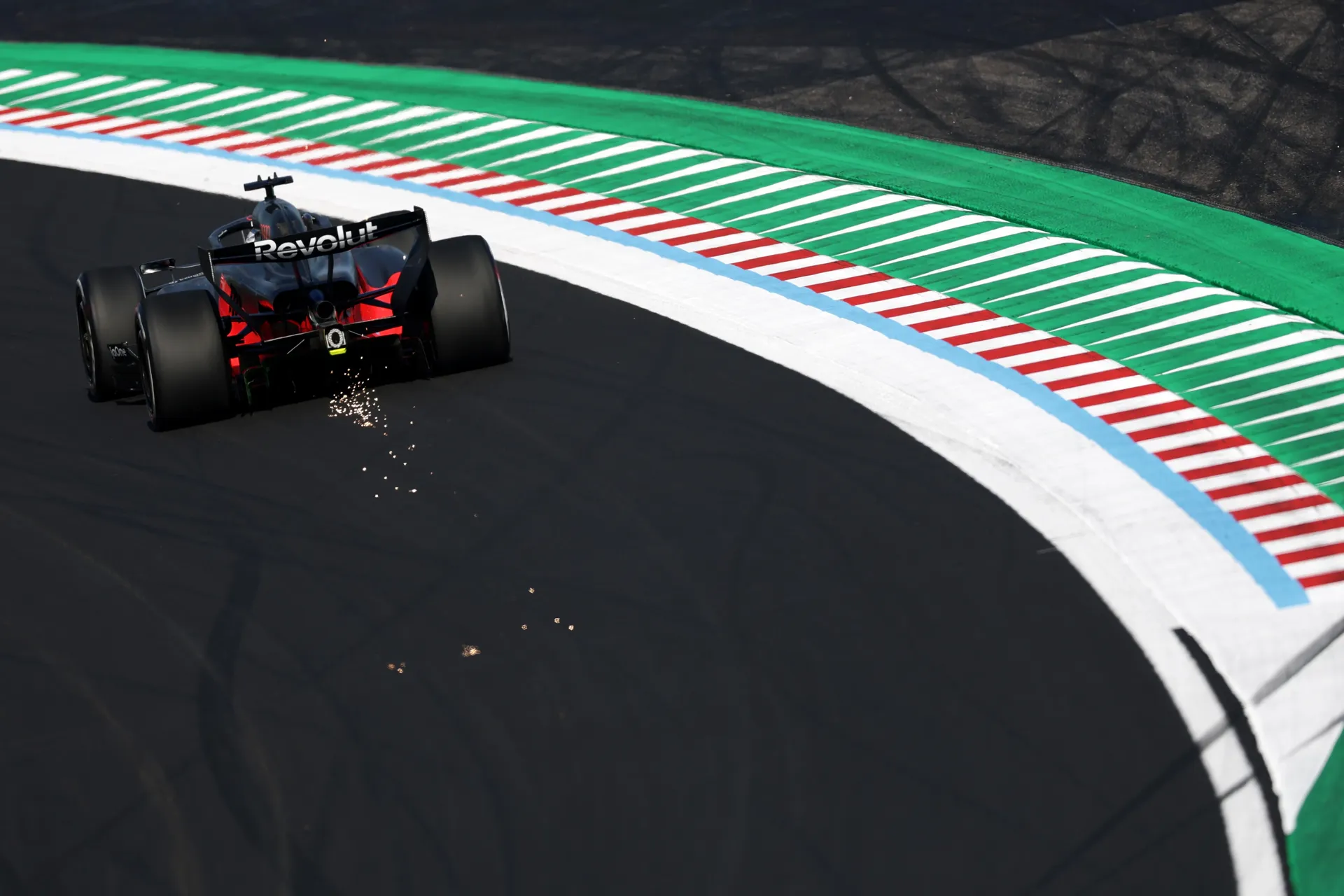 Overhead rear view of an Audi Revolut F1® Team Formula 1 car cornering at Suzuka, trailing sparks across the dark asphalt beside the striped curb.