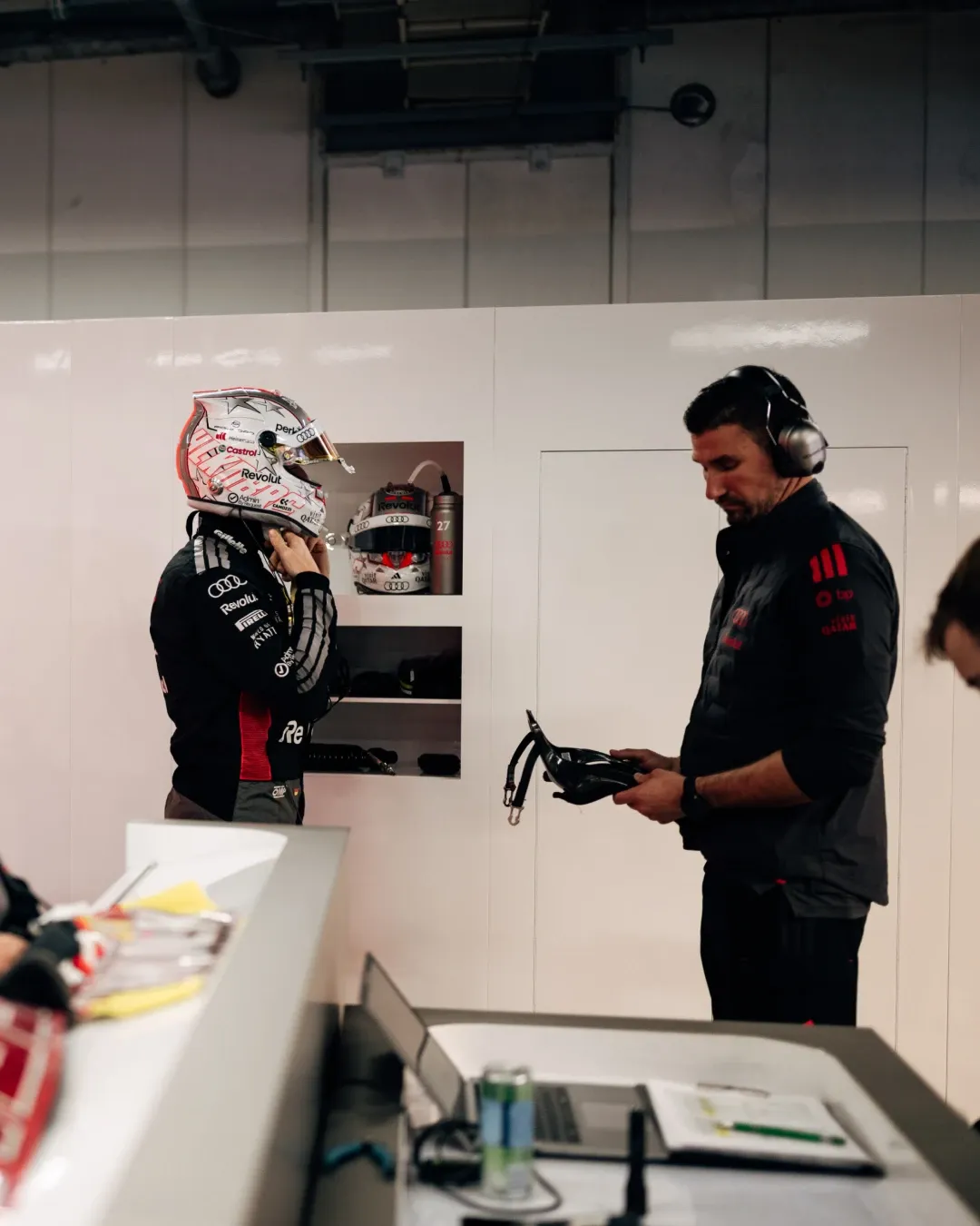 Nico Hulkenberg adjusts his helmet inside the Audi Revolut F1® Team garage during FP3 in Japan, while a team member stands opposite holding equipment, with Nico Hulkenberg’s spare helmet visible in the locker behind him.