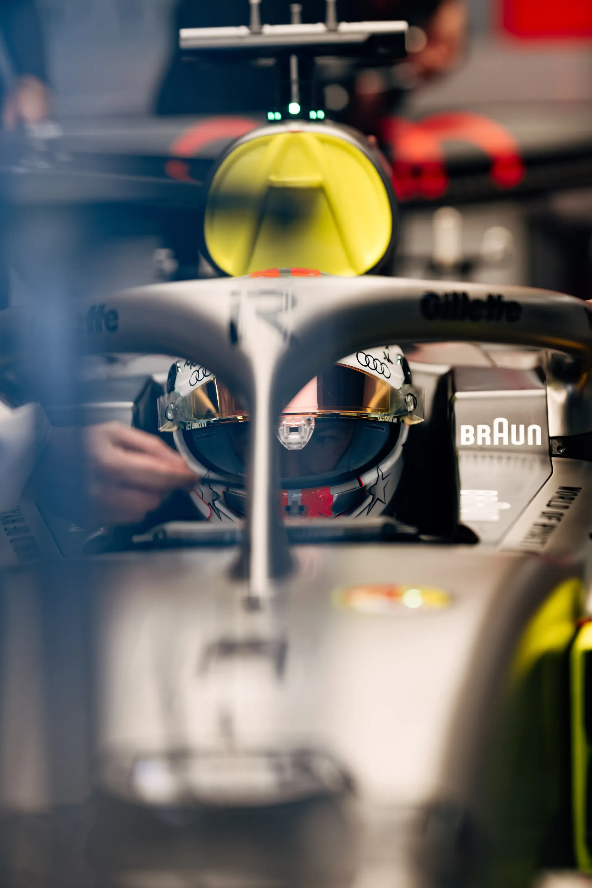 Close-up of an Audi Revolut F1® driver seated in the cockpit at Suzuka Circuit, with reflective visor and halo device in sharp focus, highlighting precision engineering, sponsor branding, and intense pre-session preparation during Japanese Grand Prix F1® media day.