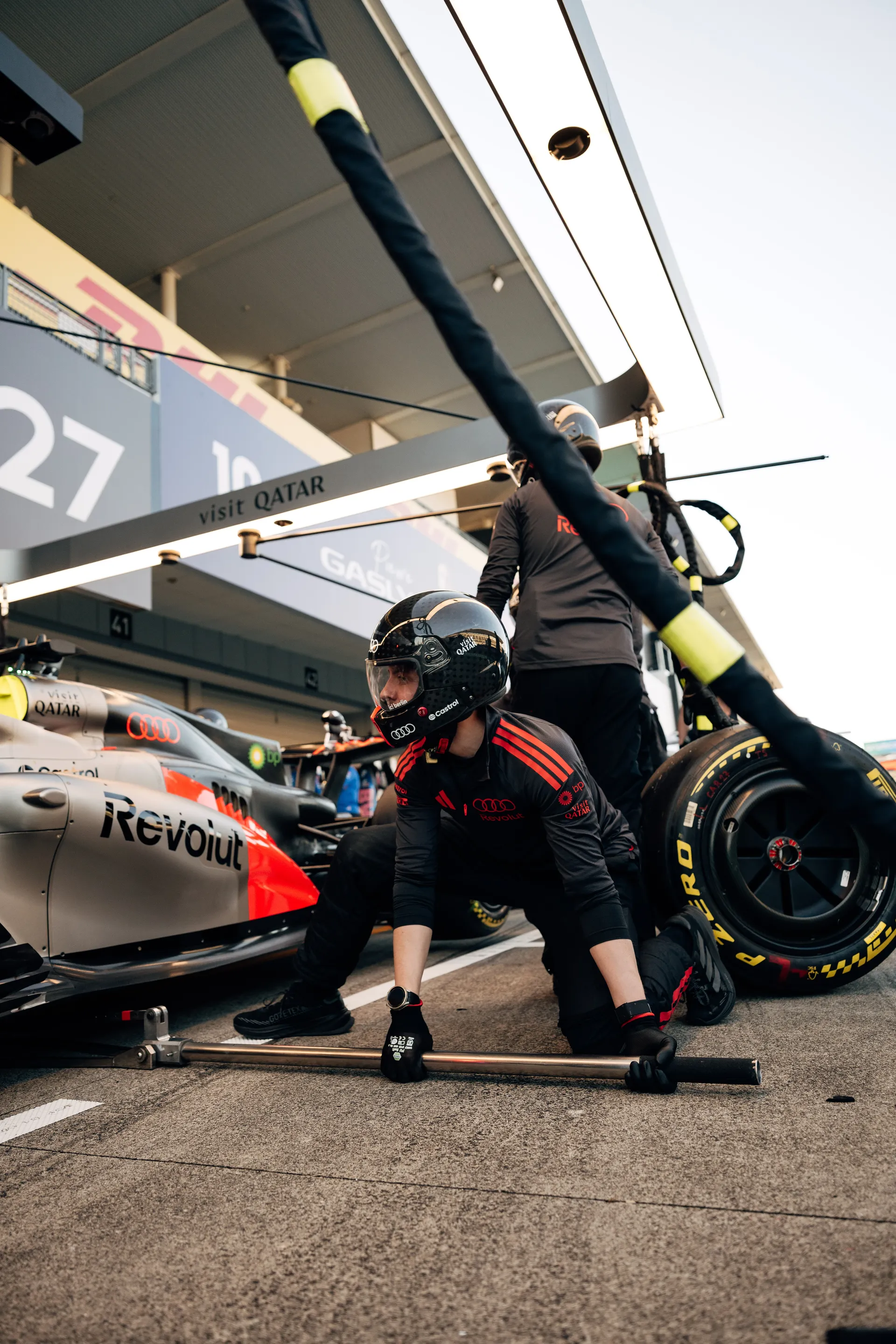 Audi Revolut F1® Team mechanic kneels beside the car in the pit lane while another crew member prepares a tyre during a pit stop setup.