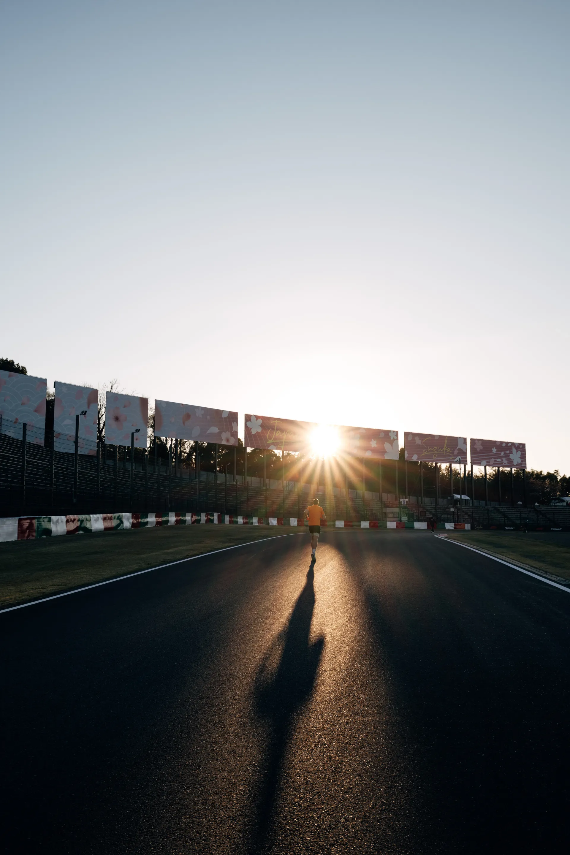 A lone runner heads toward the low sun on the Suzuka track, casting a long shadow across the asphalt.