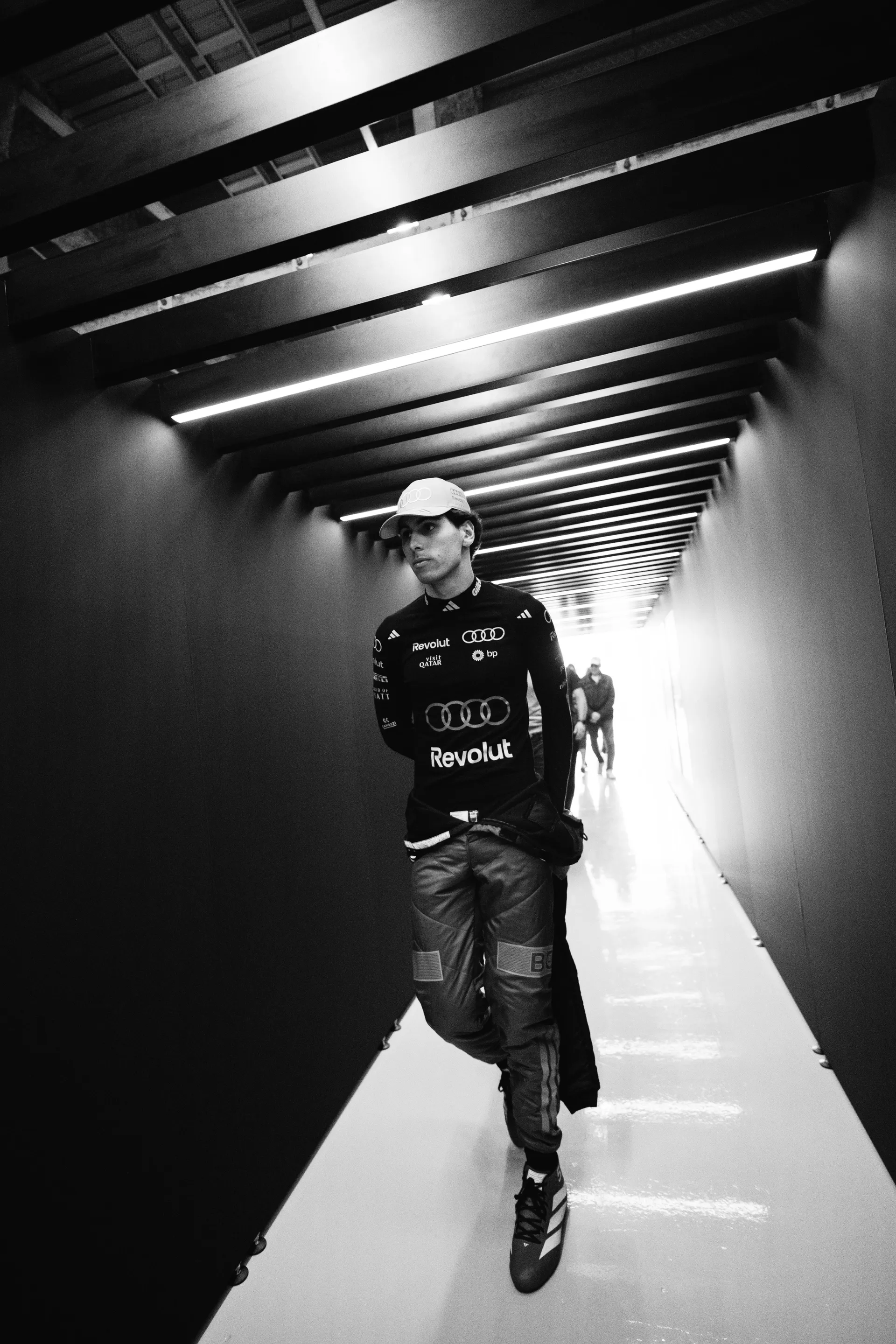 Gabriel Bortoleto walks through a narrow, black corridor in Audi Revolut F1® Team kit, lit by bright ceiling strips in a dramatic black and white portrait.