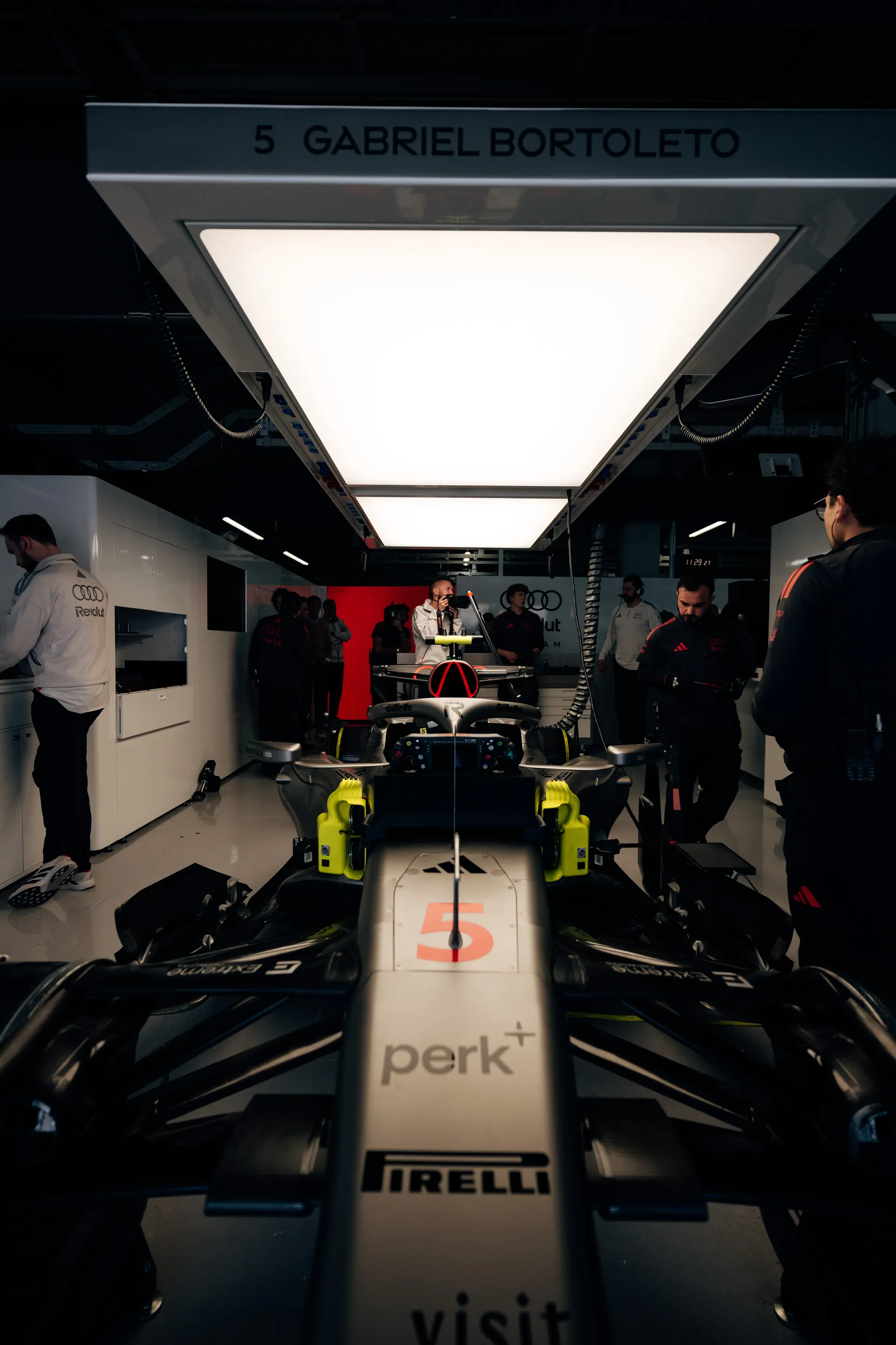 Front view of Gabriel Bortoleto’s Audi Revolut F1® Team car in the garage under a light panel marked with his name and car number 5, surrounded by crew members.