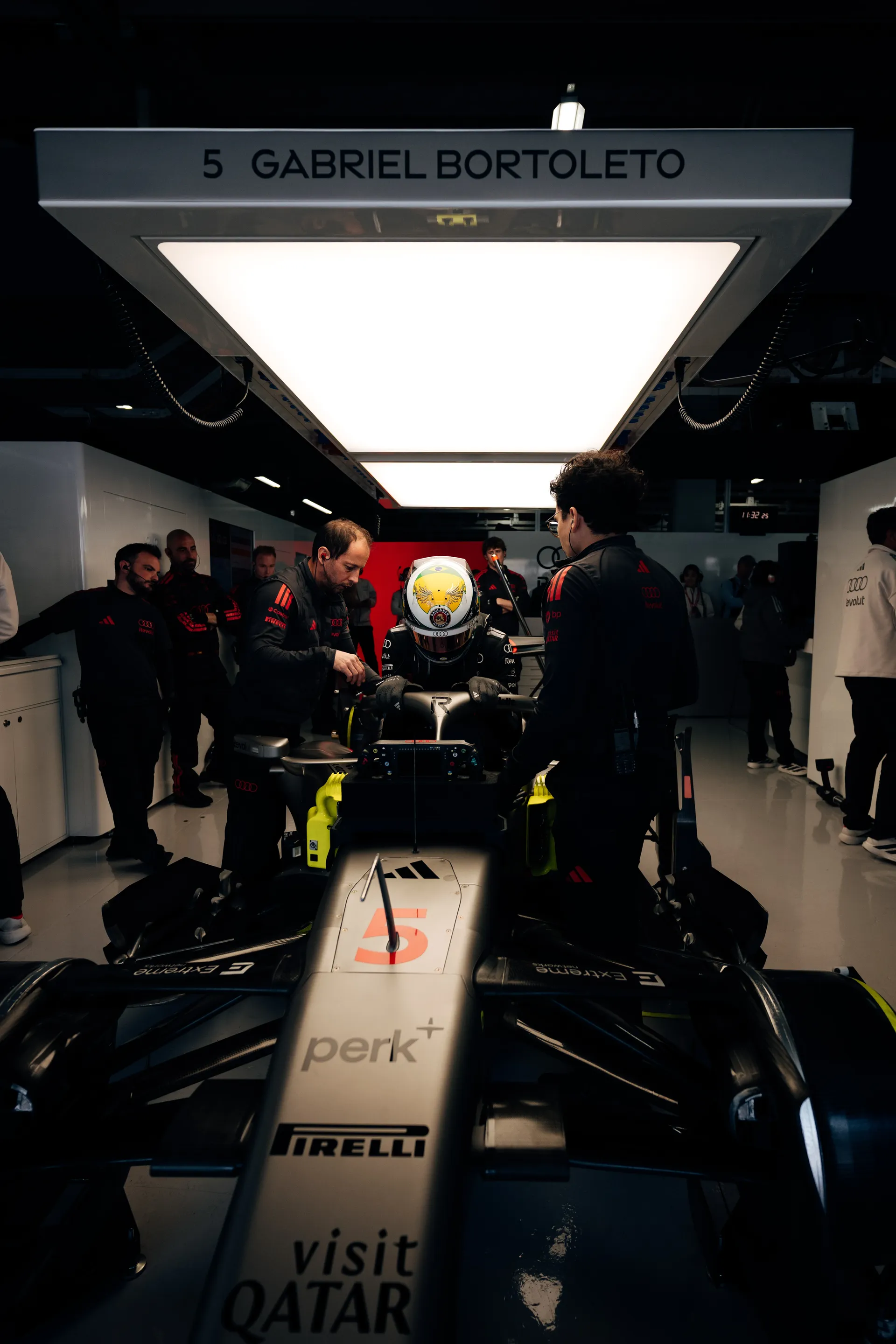 Gabriel Bortoleto sits in car number 5 in the Audi Revolut F1® Team garage while mechanics prepare the car beneath a ceiling panel labeled with his name.