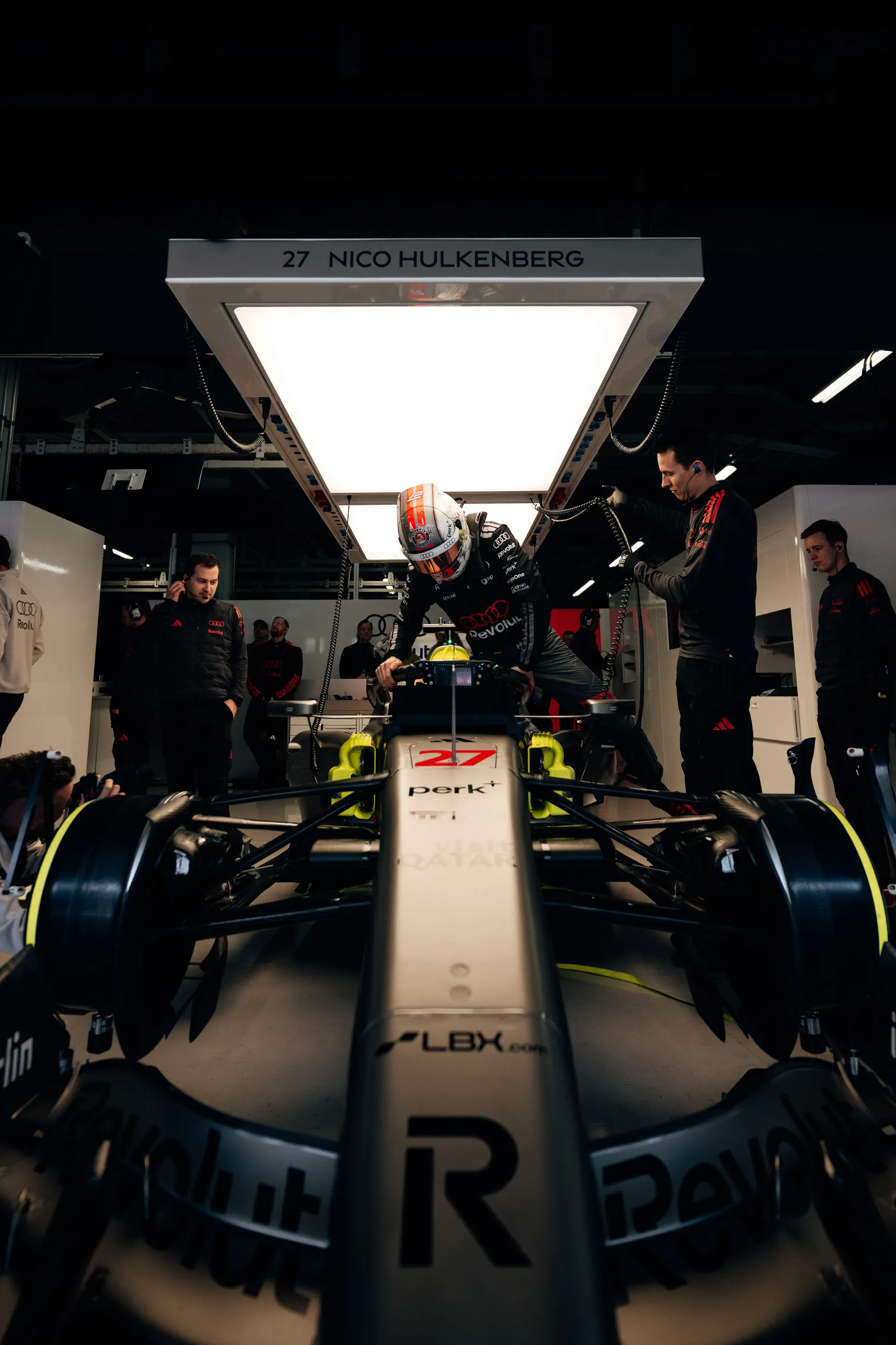 Nico Hulkenberg climbs into car number 27 in the Audi Revolut F1® Team garage as mechanics assist him under a light panel showing his name.