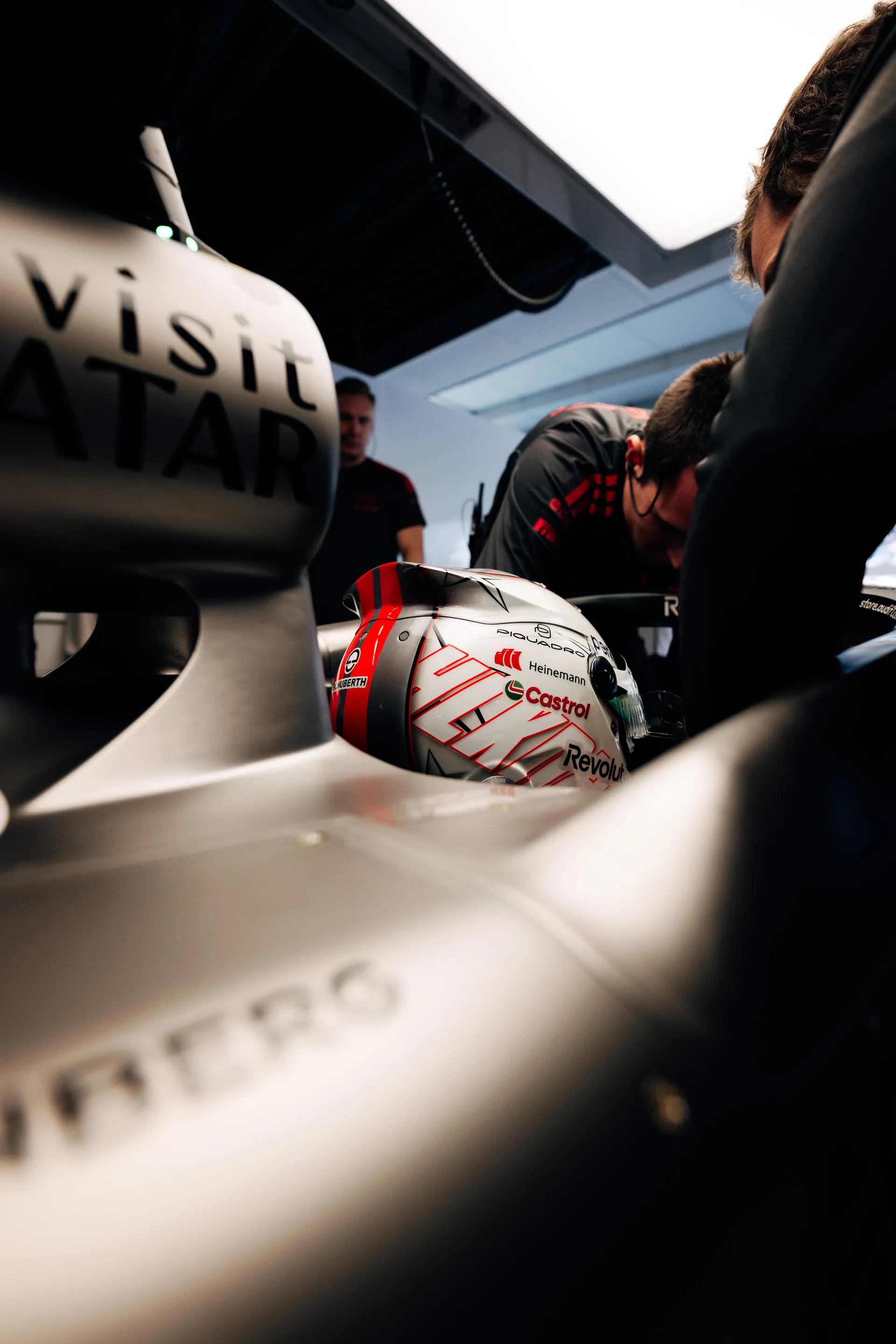 Close garage detail of Nico Hulkenberg’s helmet with red striping positioned beside the Audi Revolut F1® Team car while crew members work around it.