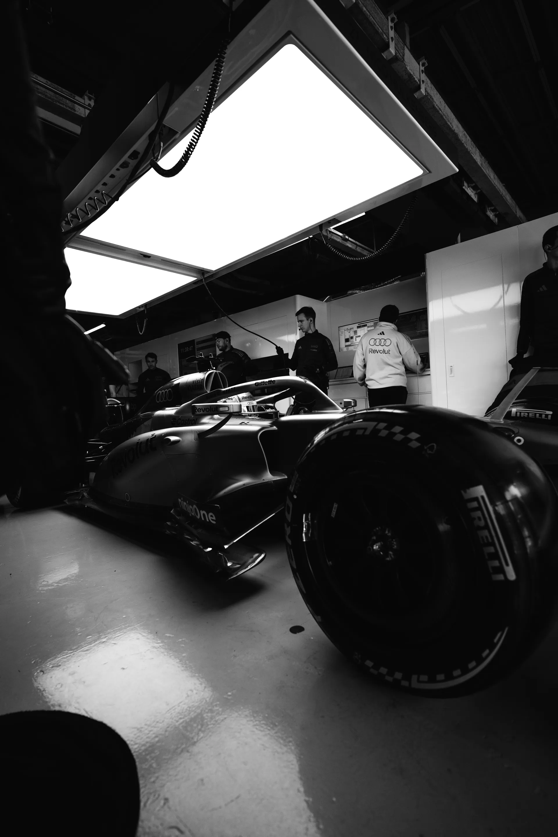 Black and white low angle shot of an Audi Revolut F1® Team car parked inside the garage under bright overhead panels, with team members working in the background.