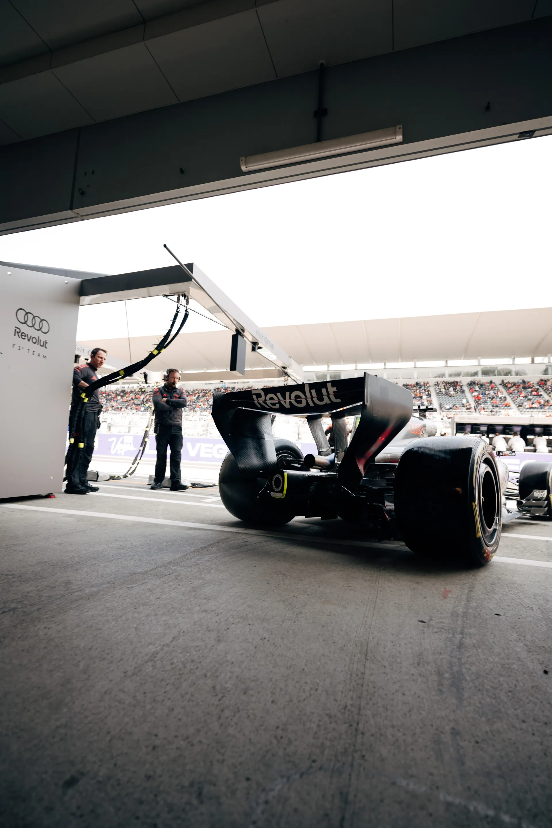 Rear three quarter view of an Audi Revolut F1® Team car waiting at the garage exit during qualifying at Suzuka, with pit crew and grandstands visible beyond.