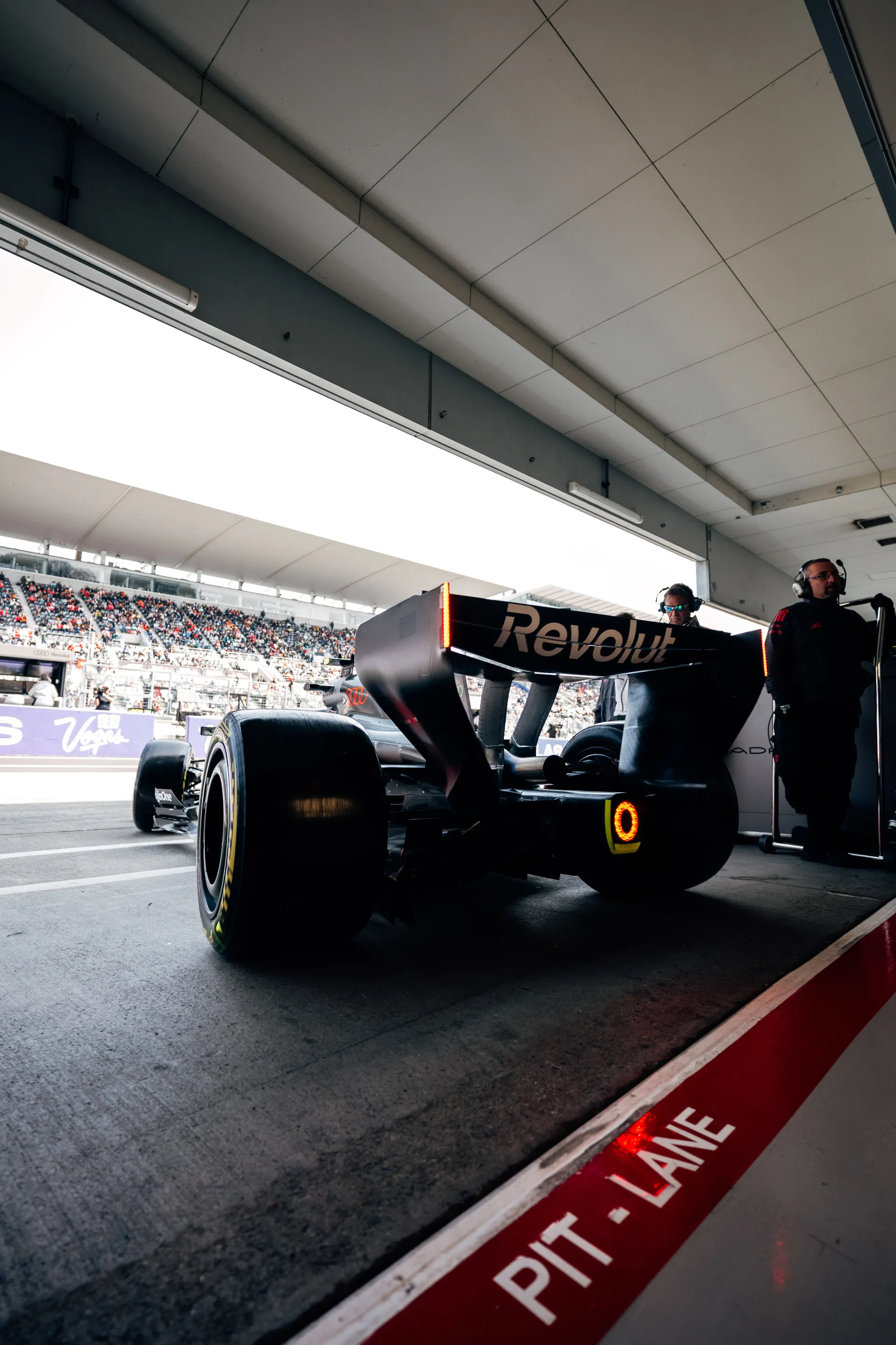 Rear view of an Audi Revolut F1® Team car at the pit lane exit with the rear light illuminated, framed by the red pit lane marking and crew members inside the garage.