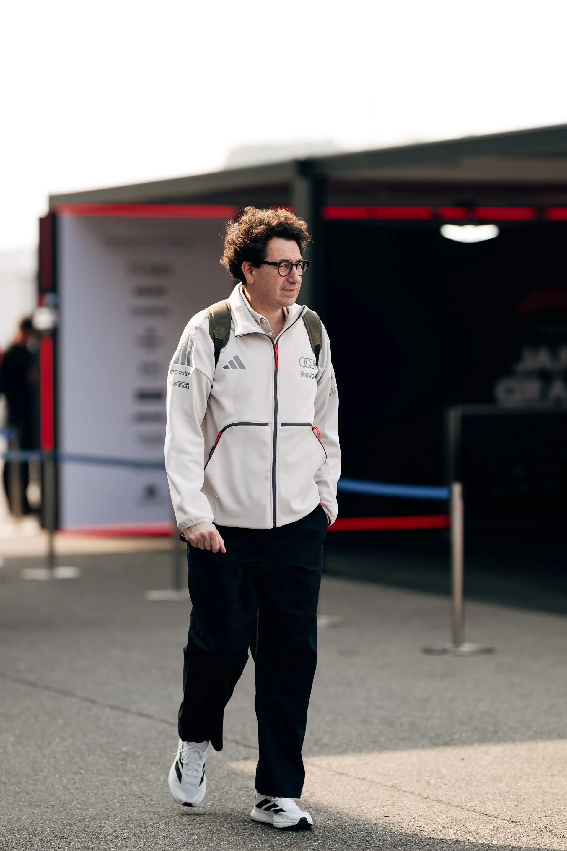 Mattia Binotto walks through the paddock in a white Audi Revolut F1® Team jacket with a backpack, passing the team hospitality area.