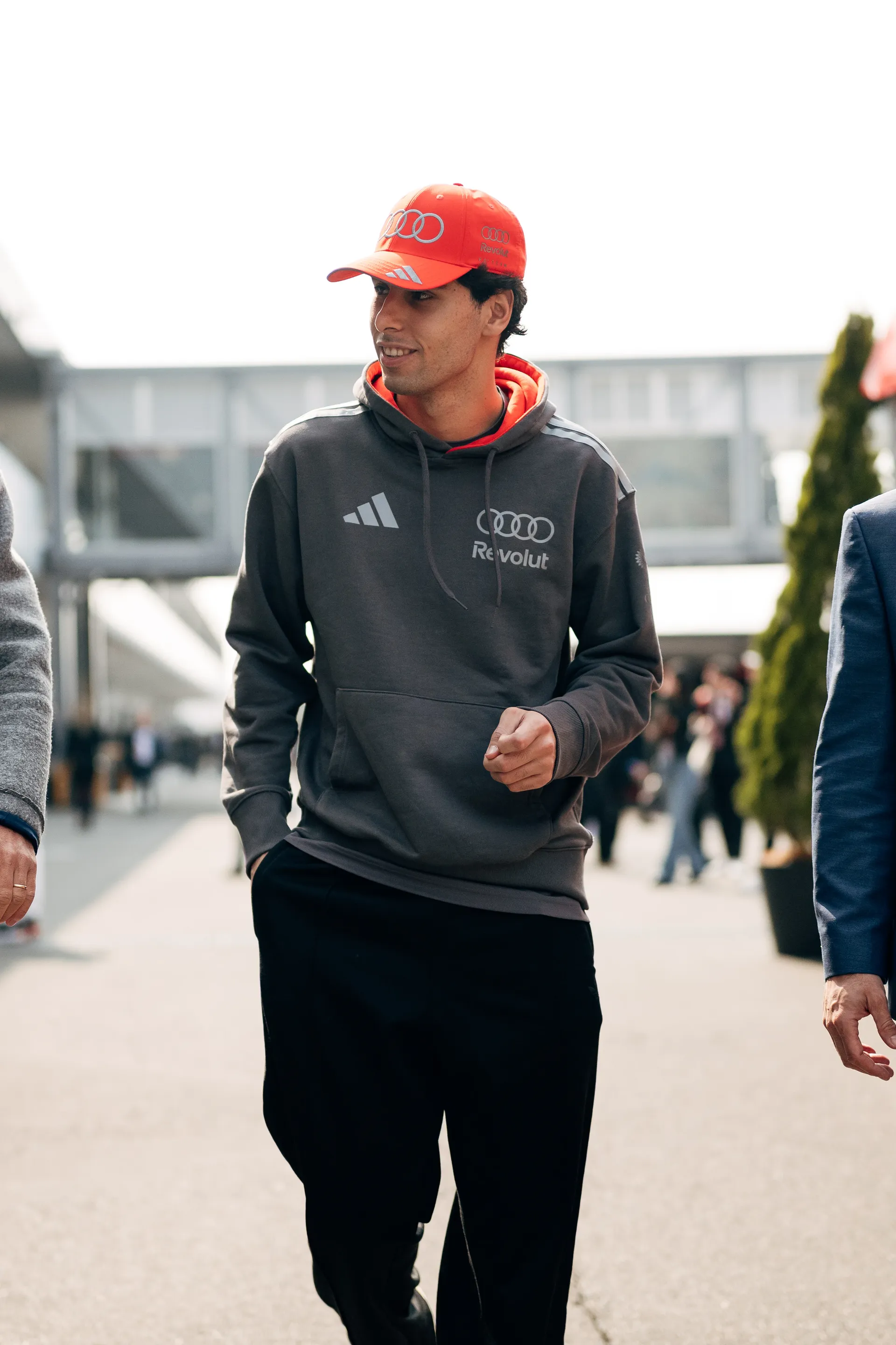 Gabriel Bortoleto walks through the paddock in a gray Audi Revolut F1® Team hoodie and red cap, smiling as he looks to the side.