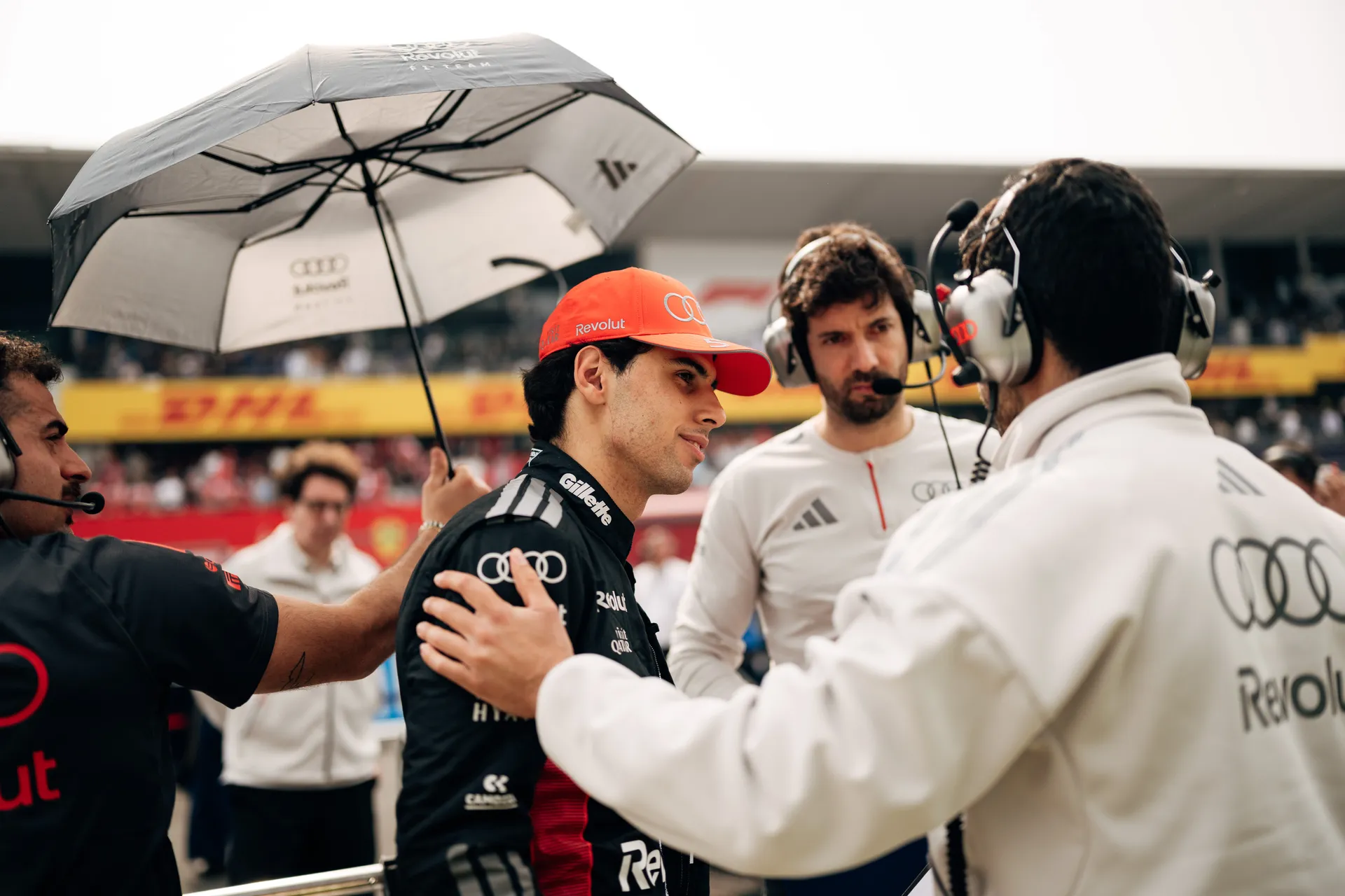 Gabriel Bortoleto speaks with Audi Revolut F1® Team staff on the grid while an umbrella is held nearby.