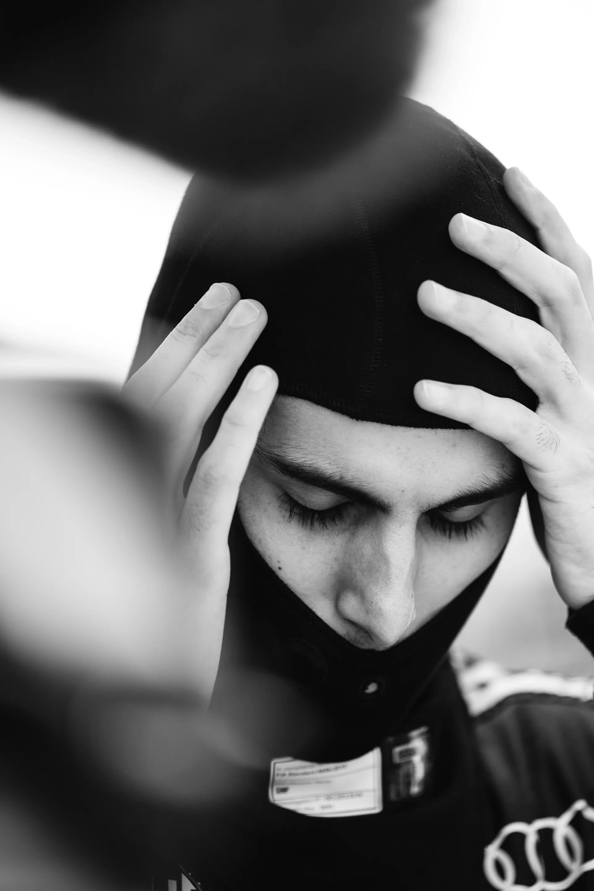 Black and white close-up of Gabriel Bortoleto of the Audi Revolut F1® Team lowering his head while adjusting his black balaclava with both hands before getting into the car.