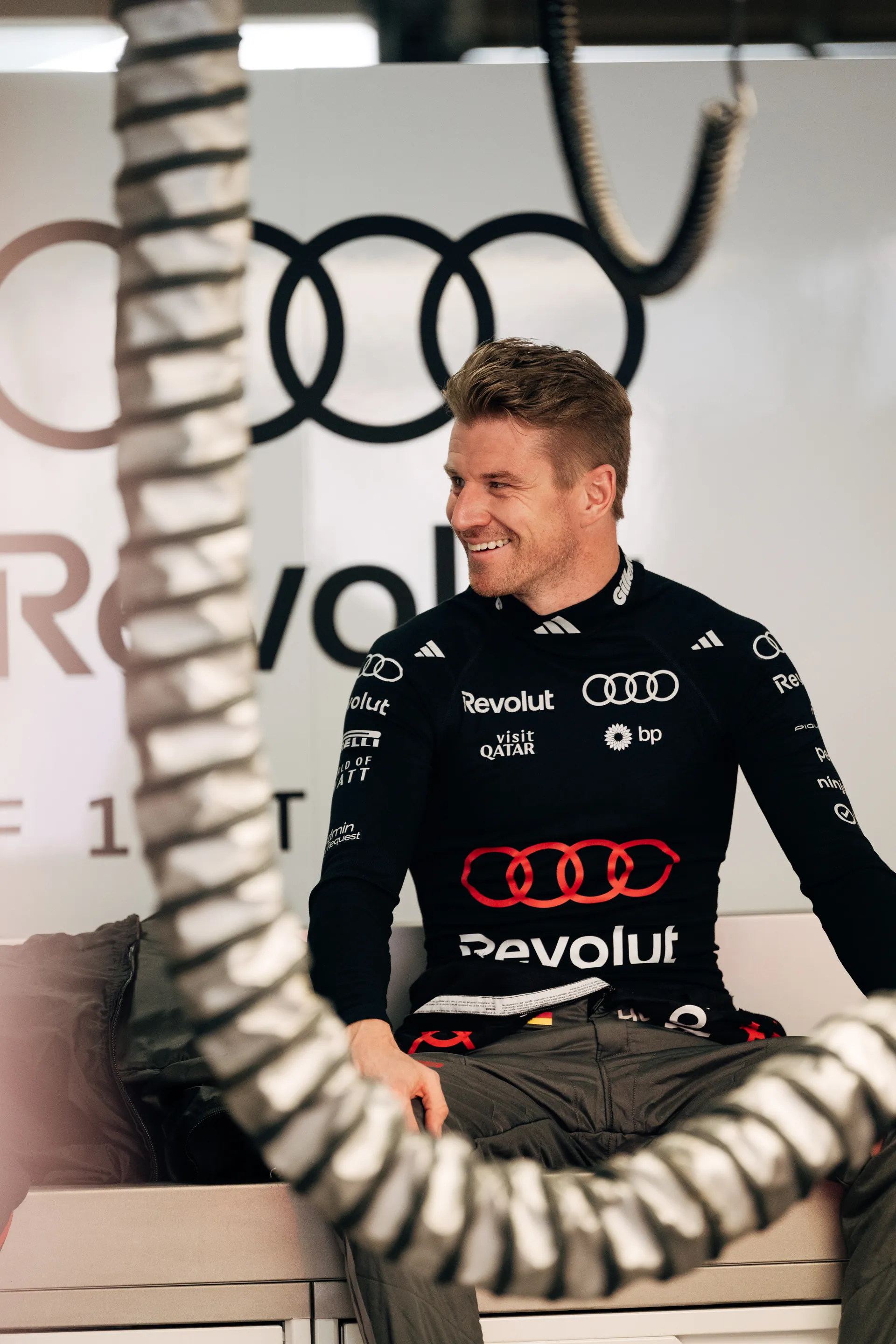 Nico Hulkenberg of Audi Revolut F1® Team sits smiling in the garage in racewear, framed by hanging garage hoses and Audi branding in the background.