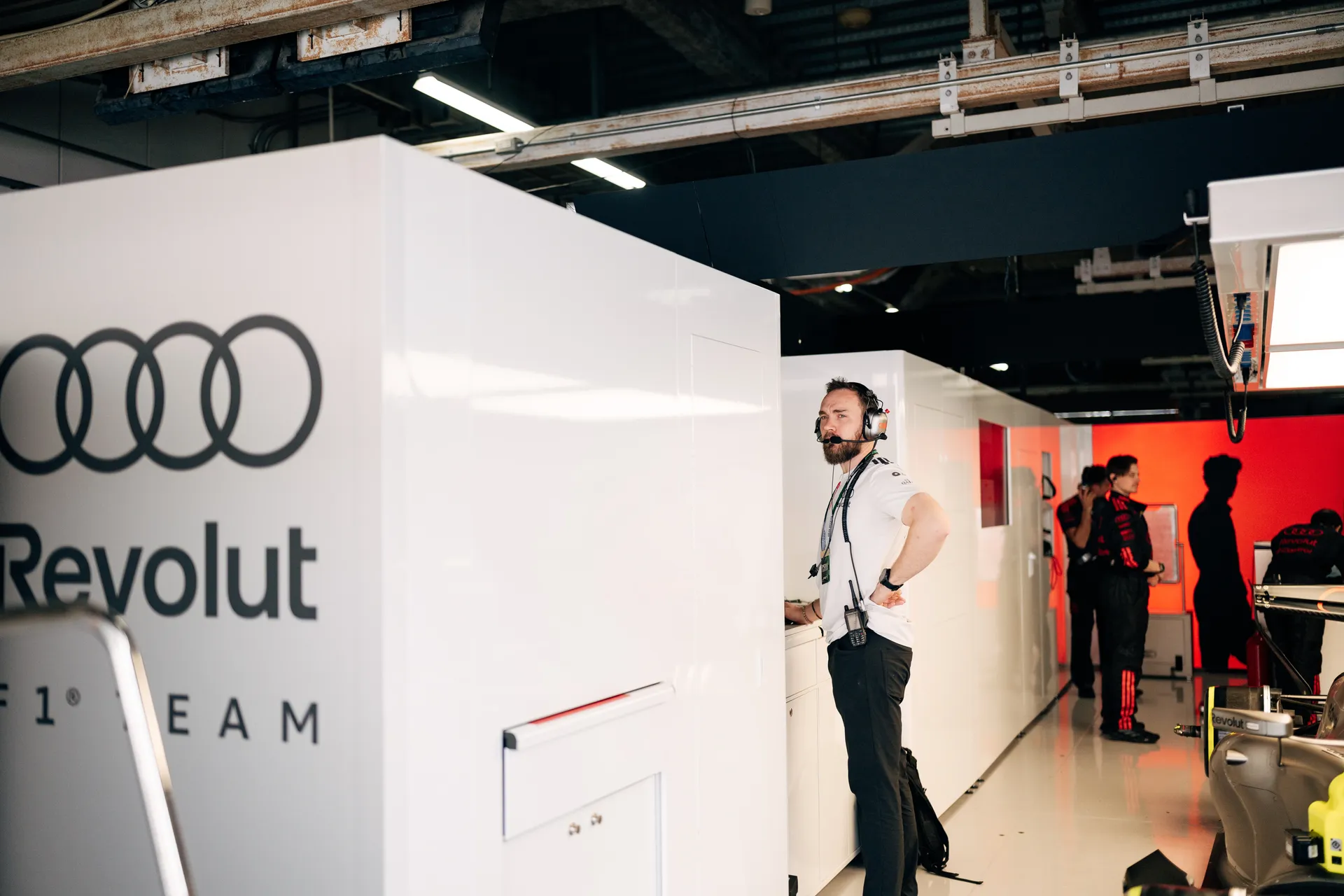 An Audi Revolut F1® Team crew member stands in the garage wearing a headset beside the white team wall, with other staff and the car visible deeper in the bay.