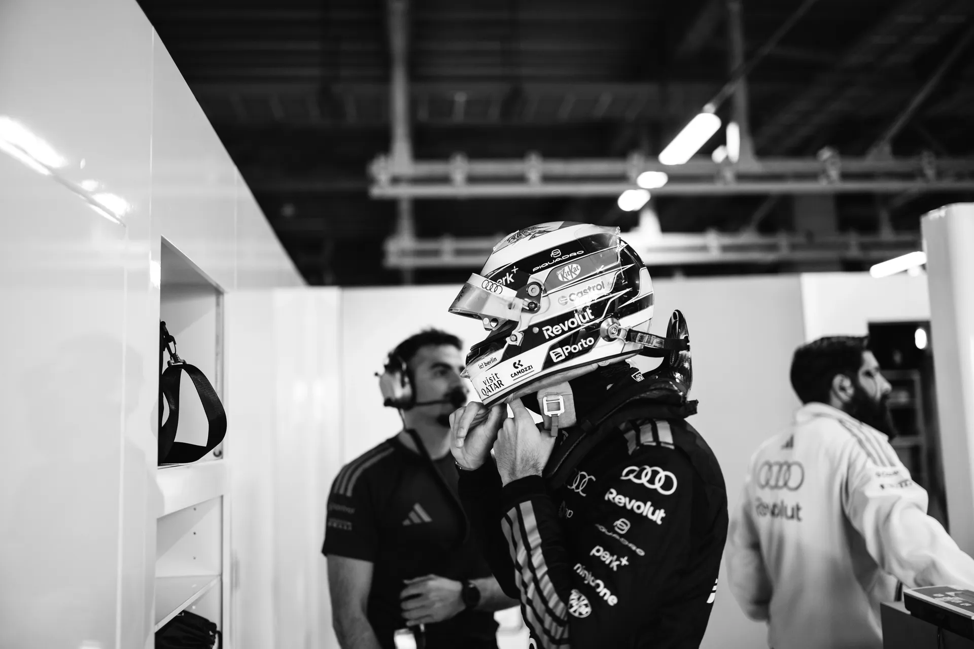 A helmeted Audi Revolut F1® Team Gabriel Bortoleto fastens his helmet strap in the garage while team staff stand nearby in the background.