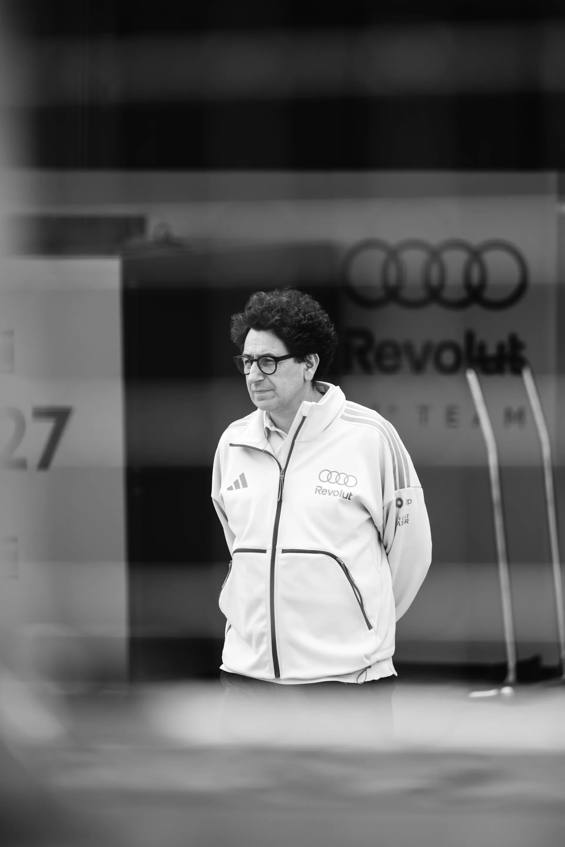 Mattia Binotto of Audi Revolut F1® Team stands with his hands behind his back outside the garage, photographed in black and white with the team branding behind him.