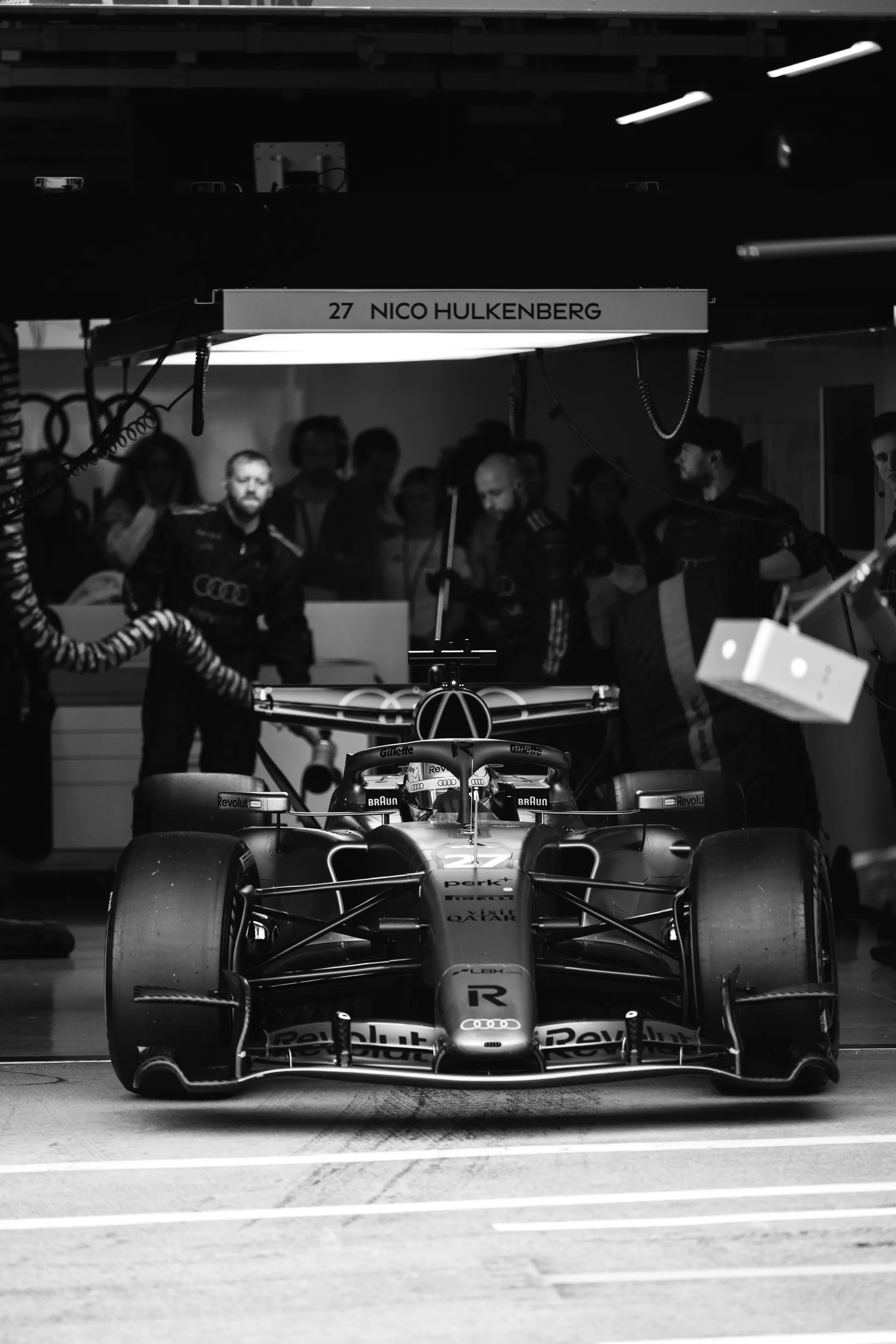 Nico Hulkenberg’s Audi Revolut F1® Team car waits at the garage exit beneath a sign marked with his name and number 27, shown in black and white.