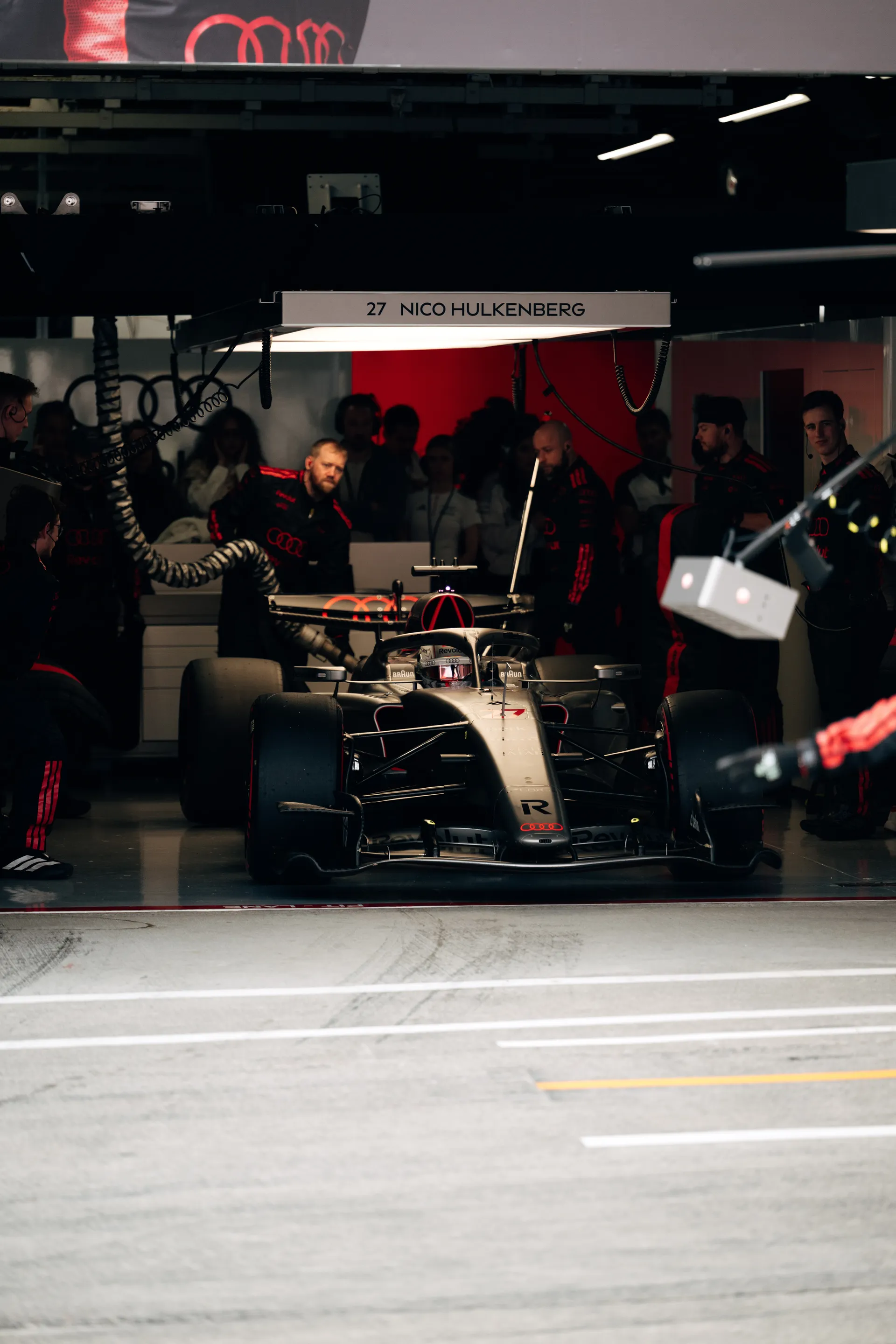Nico Hulkenberg’s Audi Revolut F1® Team car sits at the garage exit under the 27 Nico Hulkenberg nameplate while crew members watch from the shadows behind.