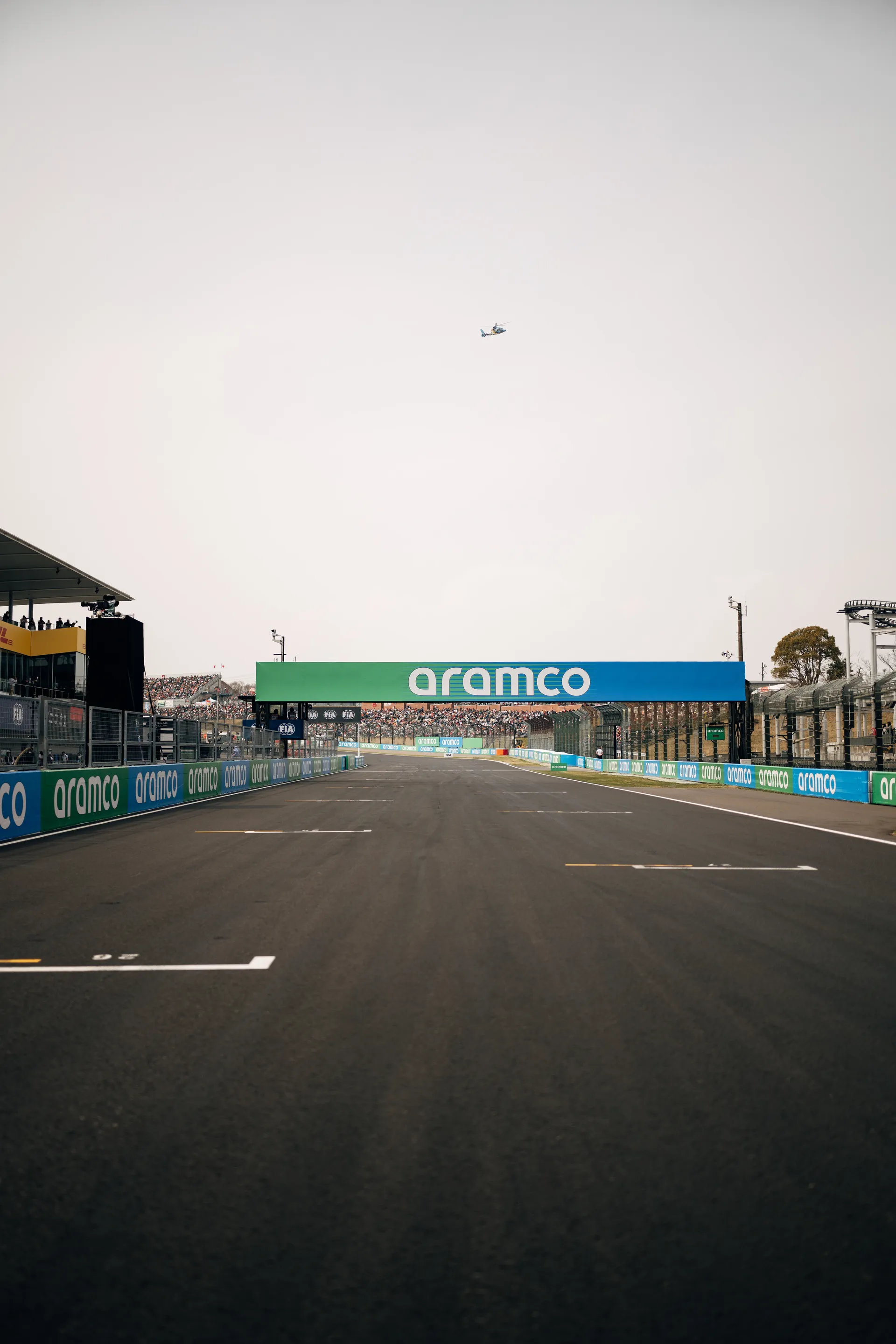 An empty Suzuka start finish straight stretches toward an Aramco bridge, with grandstands on both sides and a helicopter high above the circuit.