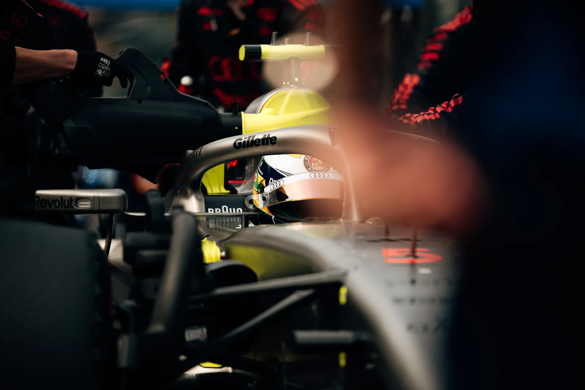 Gabriel Bortoleto sits in his Audi Revolut F1® Team car in the garage, seen through the cockpit and blurred team members, with his helmet visible behind the halo.