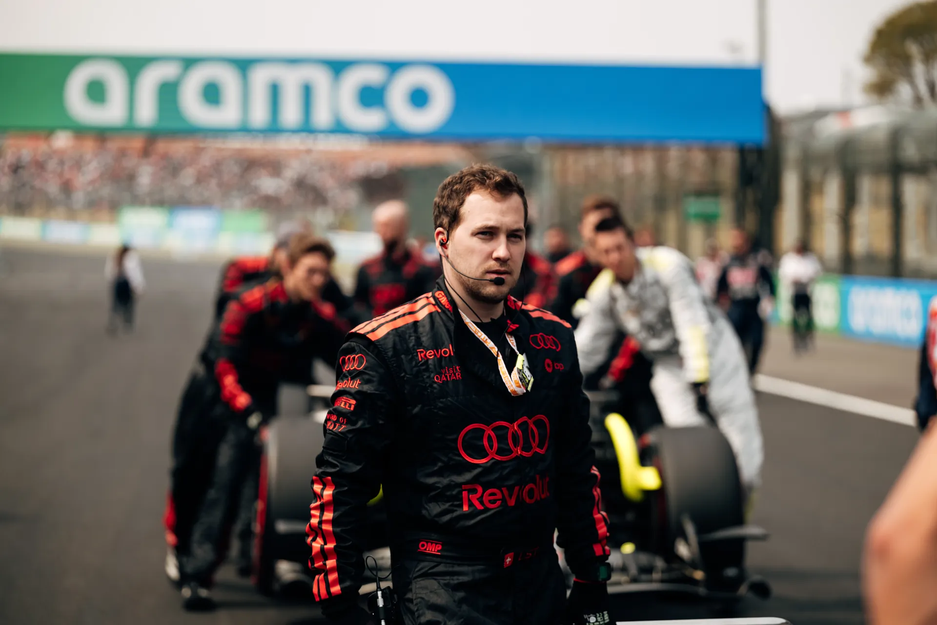 A focused Audi Revolut F1® Team crew member wearing a headset walks ahead of Gabriel Bortoleto’s car as the team moves along the grid.