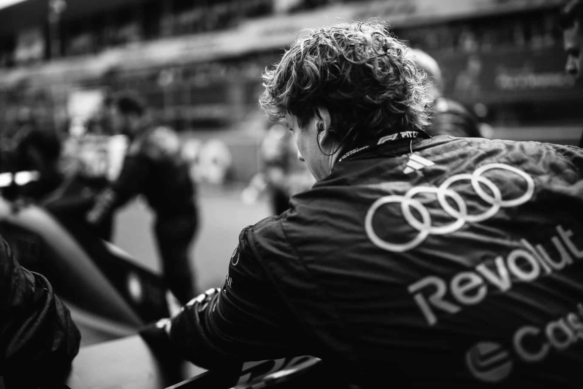 A crew member from Audi Revolut F1® Team leans over the car on the grid in black and white, with blurred team activity in the background.