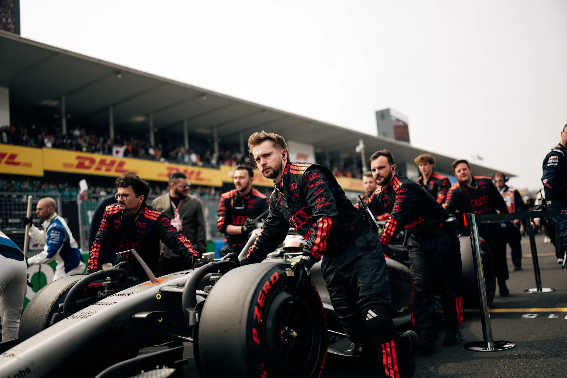 Several Audi Revolut F1® Team crew members push the car along the Suzuka grid, with one mechanic leading at the front and grandstands full of spectators behind them.