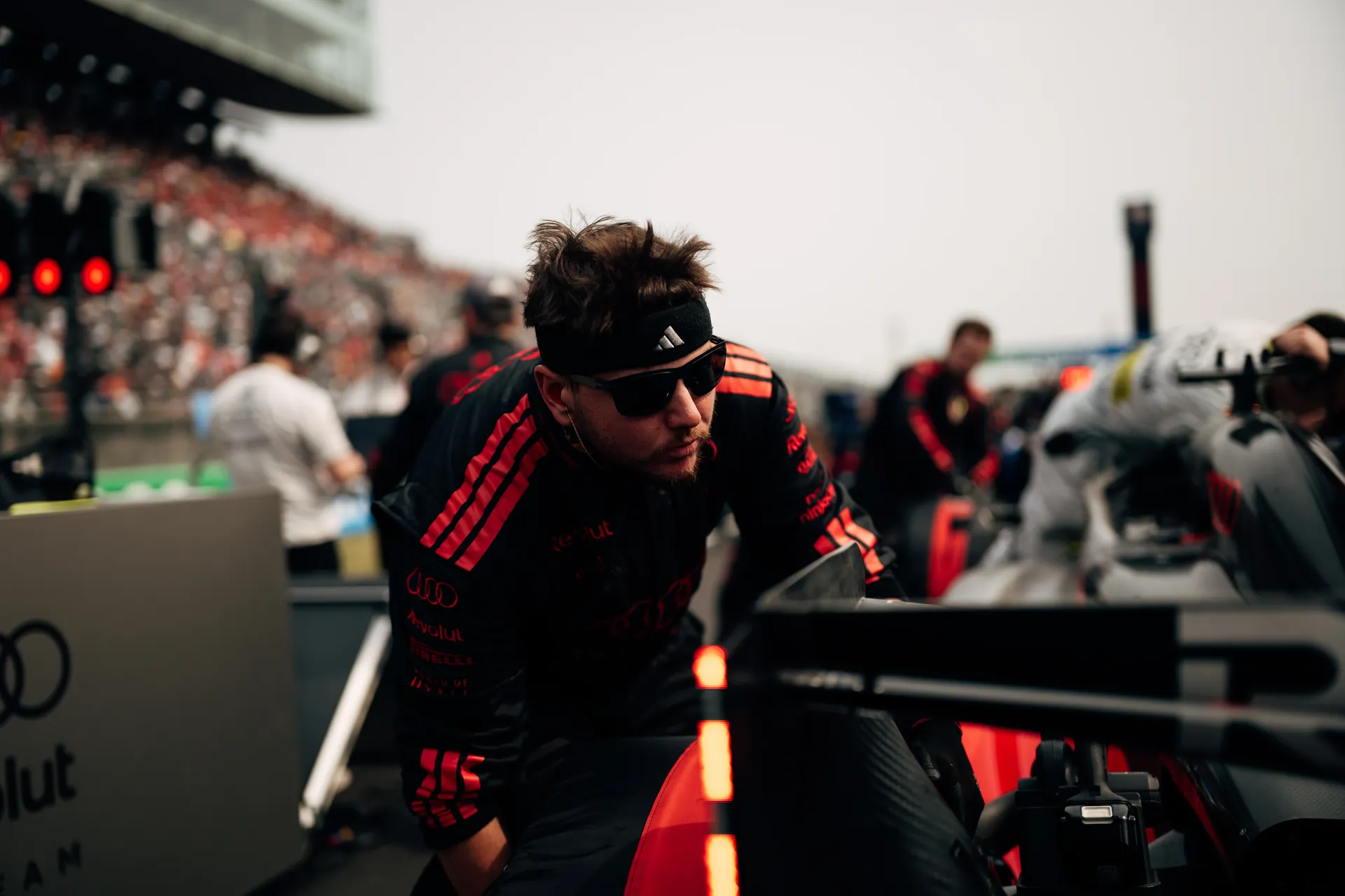 An Audi Revolut F1® Team mechanic wearing sunglasses and a headband leans over the car on the grid, with the rear light and blurred pit lane activity behind him.