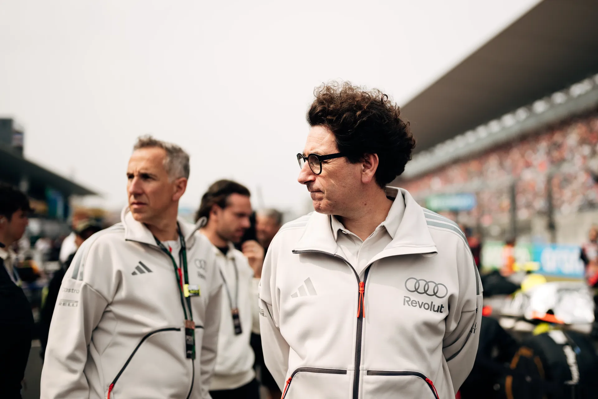 Mattia Binotto of Audi Revolut F1® Team stand on the Suzuka grid in white team jackets, looking across the track with the grandstands blurred behind them.