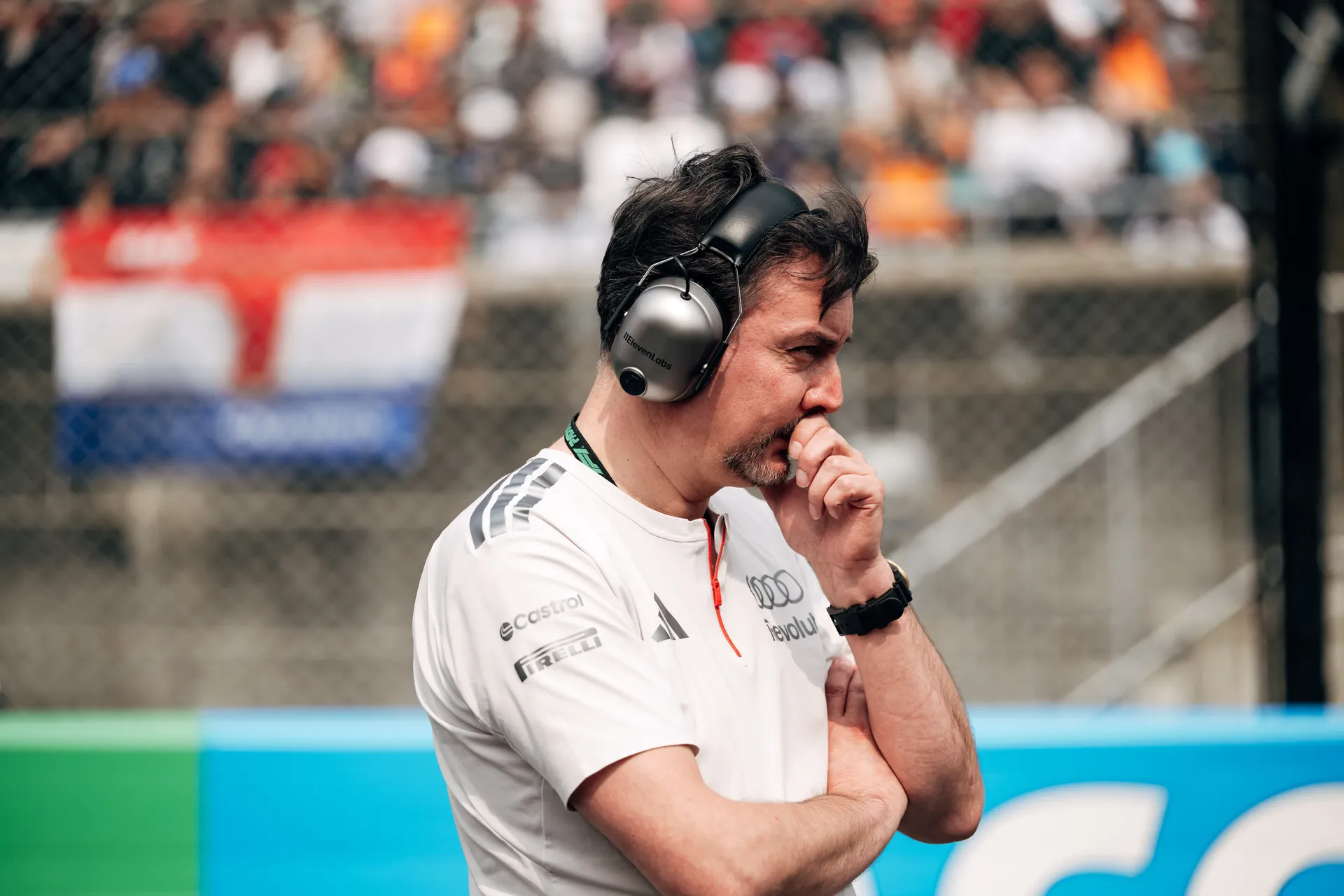 Audi Revolut F1® Team member James Key wearing headphones stands at the side of the grid in profile, watching the track with a crowded grandstand in the background.