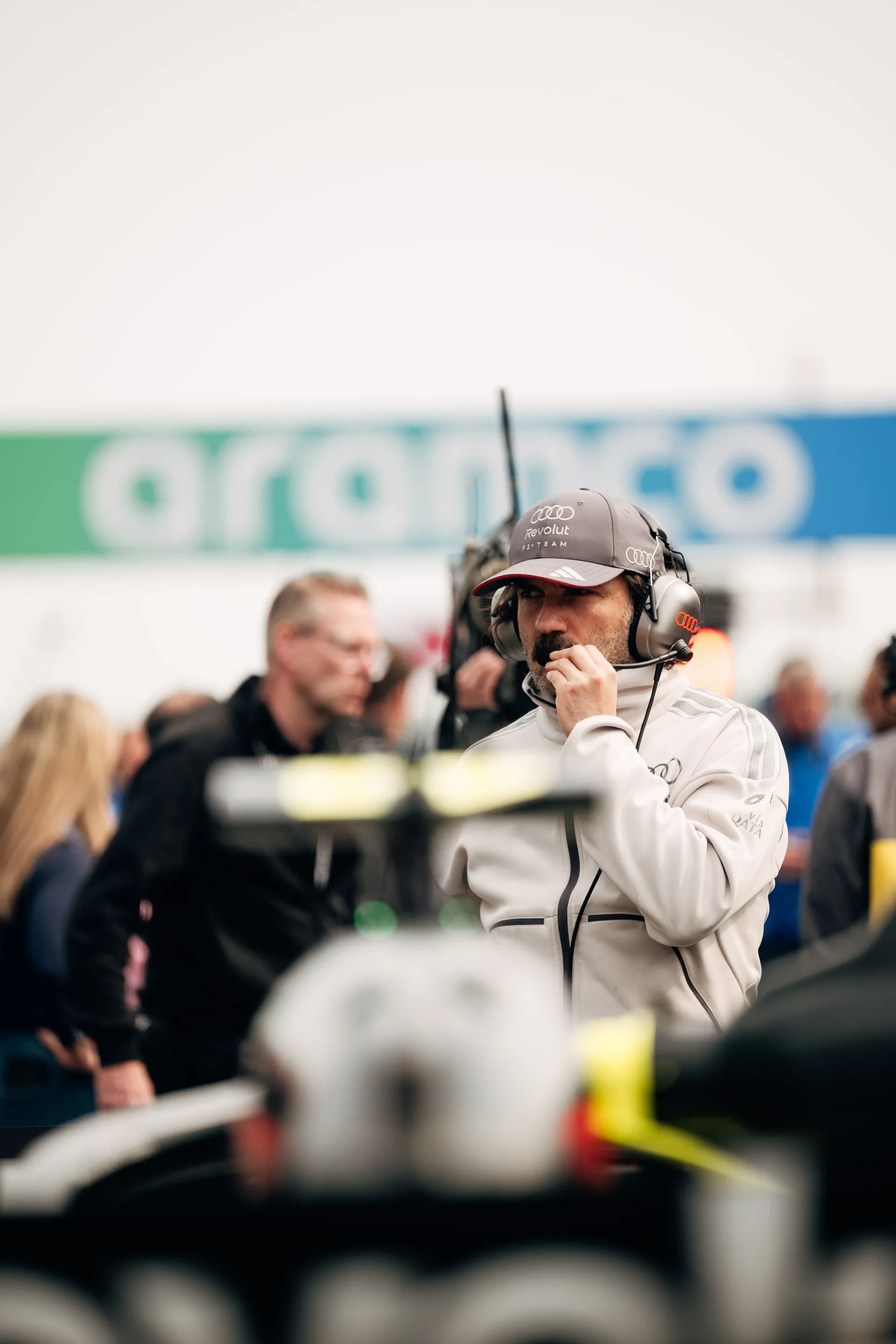 An Audi Revolut F1® Team engineer in a team cap and headset stands near the car on the grid, speaking into his radio with Aramco signage blurred in the background.