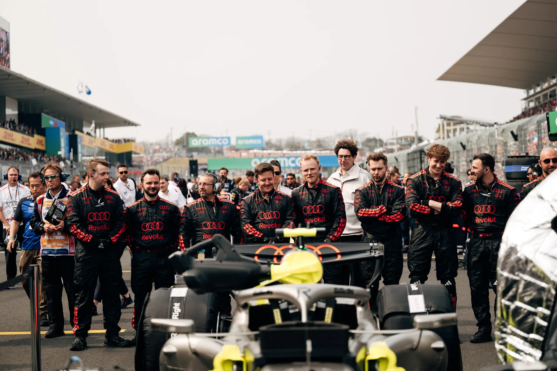 Audi Revolut F1® Team crew members gather behind the car on the Suzuka grid, with Mattia Binotto visible among the group as they watch preparations unfold.
