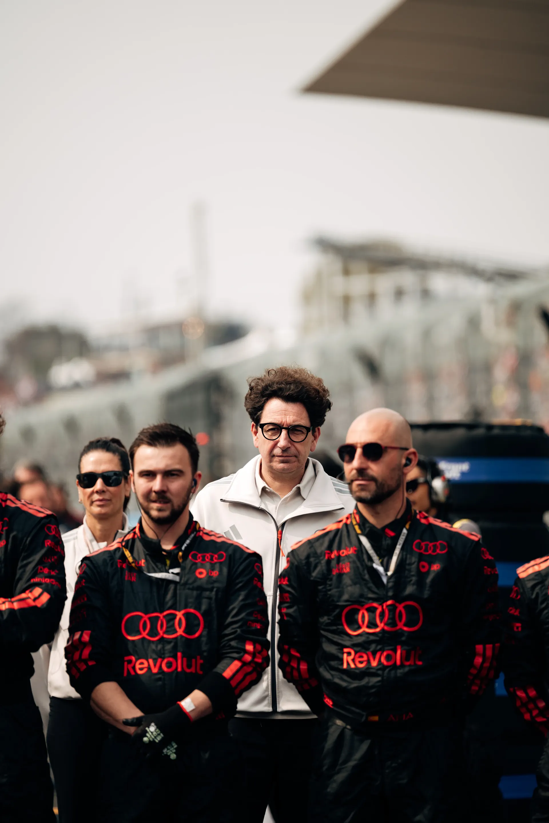Mattia Binotto of Audi Revolut F1® Team stands behind crew members on the grid, framed between team staff in black racewear.