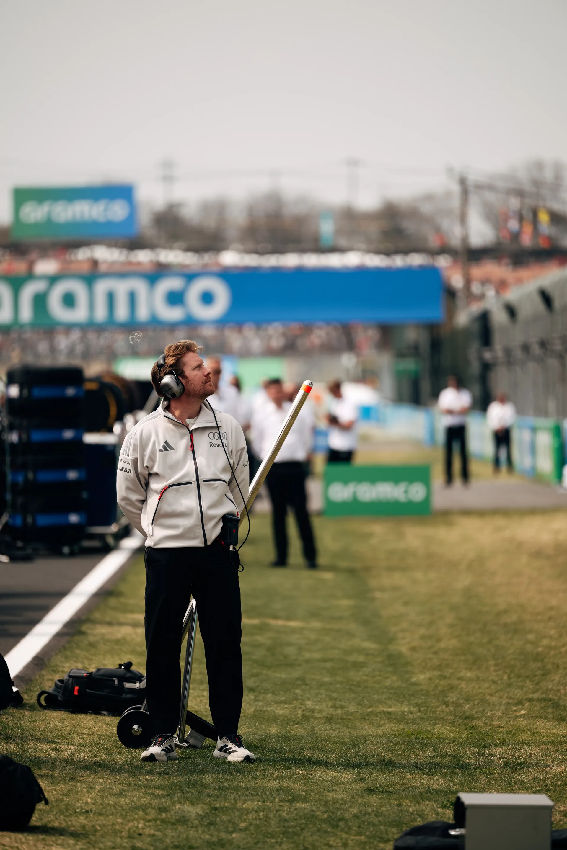 Team member of Audi Revolut F1® Team stands on the grass beside the track with a headset on, looking upward while holding a long measuring wand near the Aramco bridge.