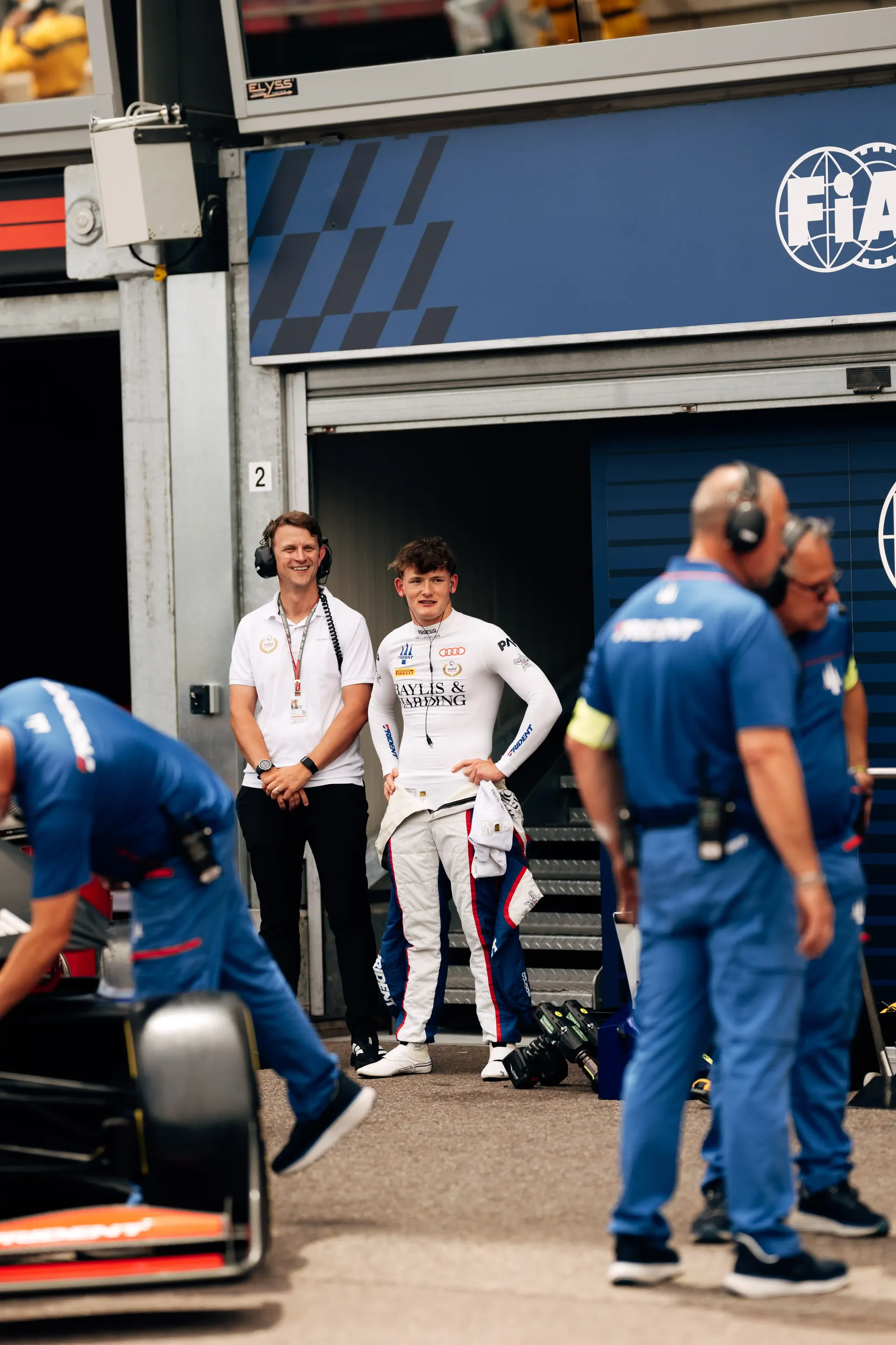 Freddie Slater stands outside the Formula 3 garage alongside a team member in the Monaco paddock before a track session.