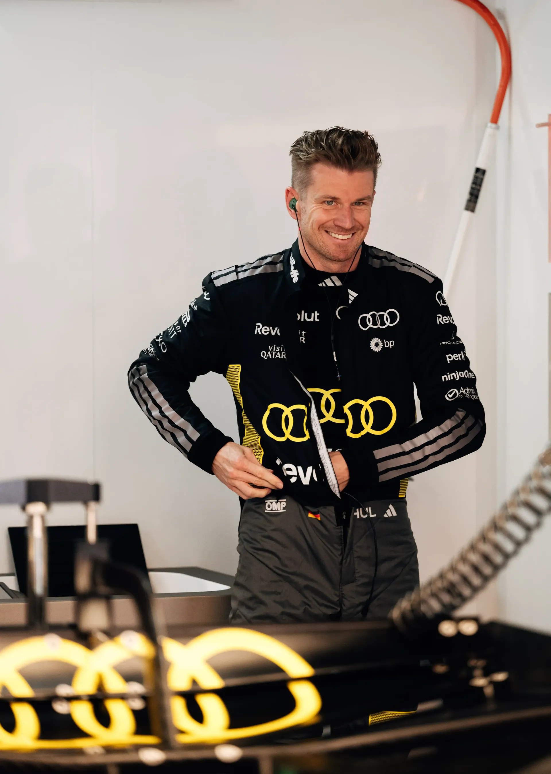 Nico Hülkenberg stands smiling beside the Audi Revolut F1® Team car R26 inside the garage.