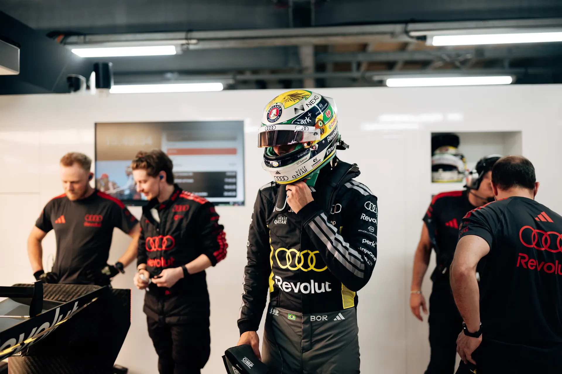 Audi Revolut F1® Team driver Gabriel Bortoleto wearing a helmet stands with engineers and mechanics inside the garage.