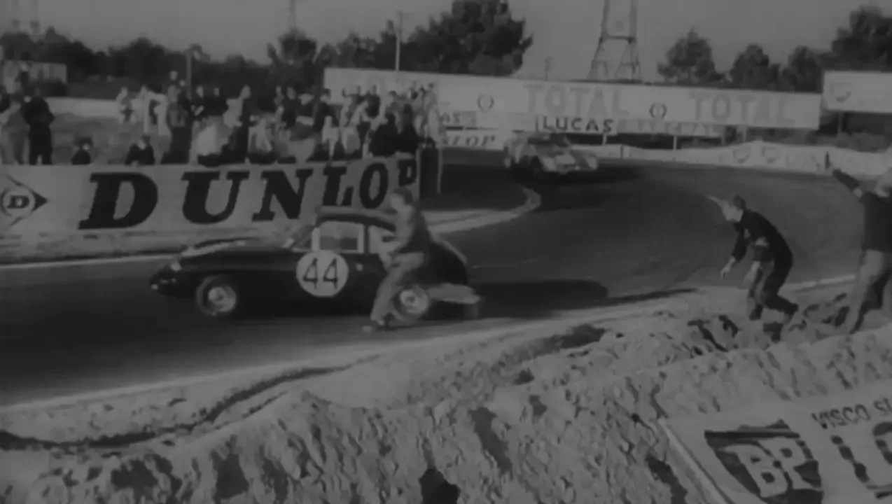 Black and white archive footage of vintage sports cars racing past a Dunlop bridge at the track