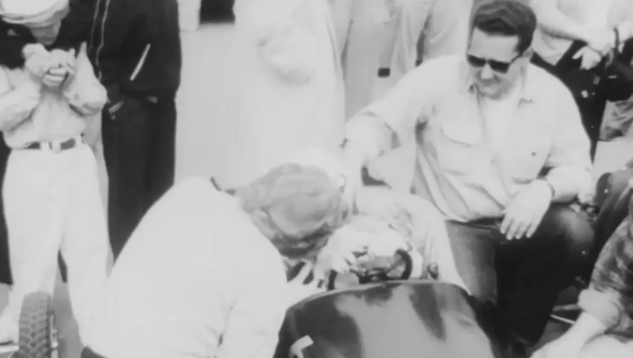 Black and white archive footage of a young driver in a race car surrounded by people