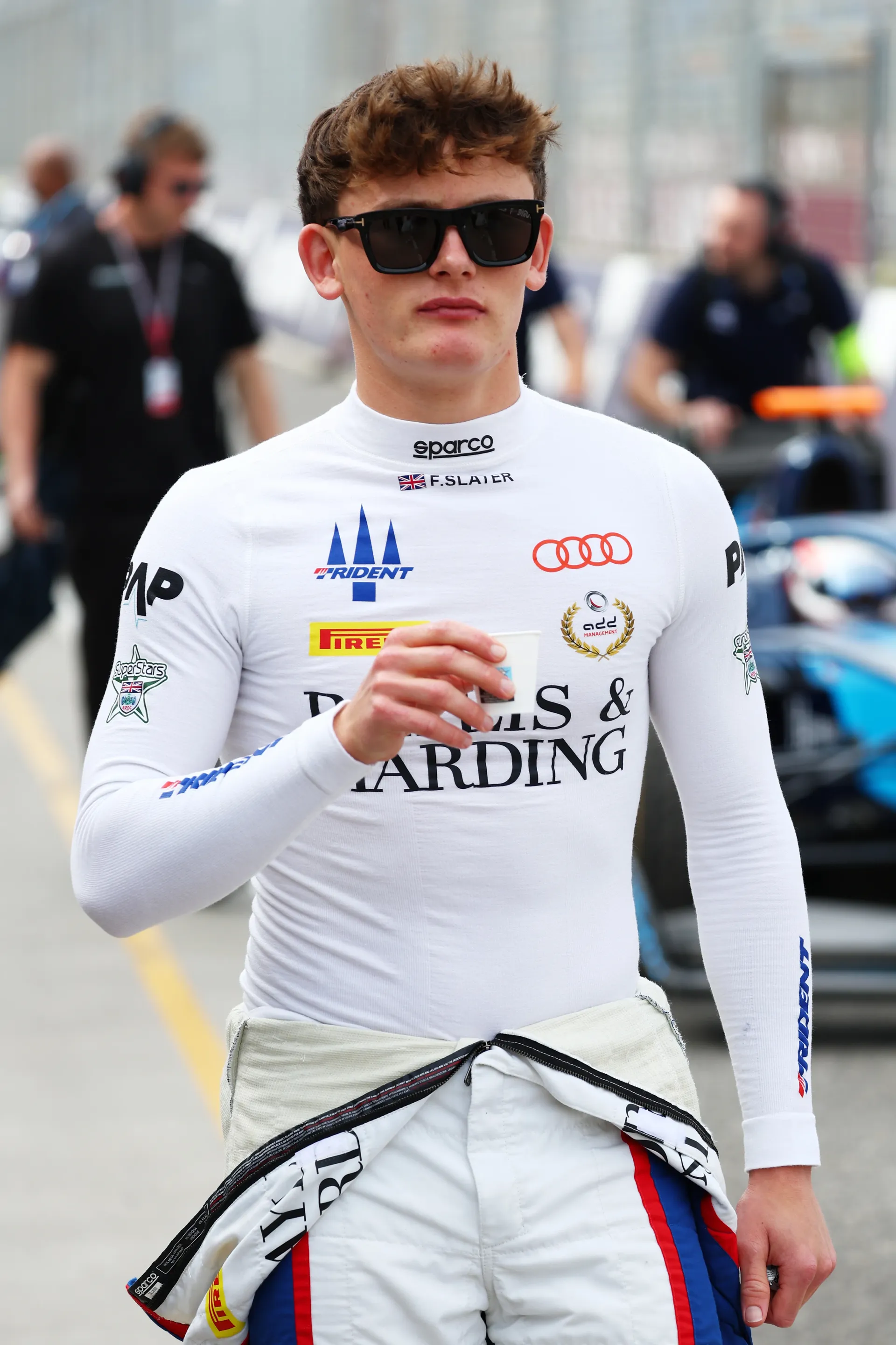 Freddie Slater walking through the Formula 3 pit lane wearing sunglasses and race base layer holding a cup during race weekend